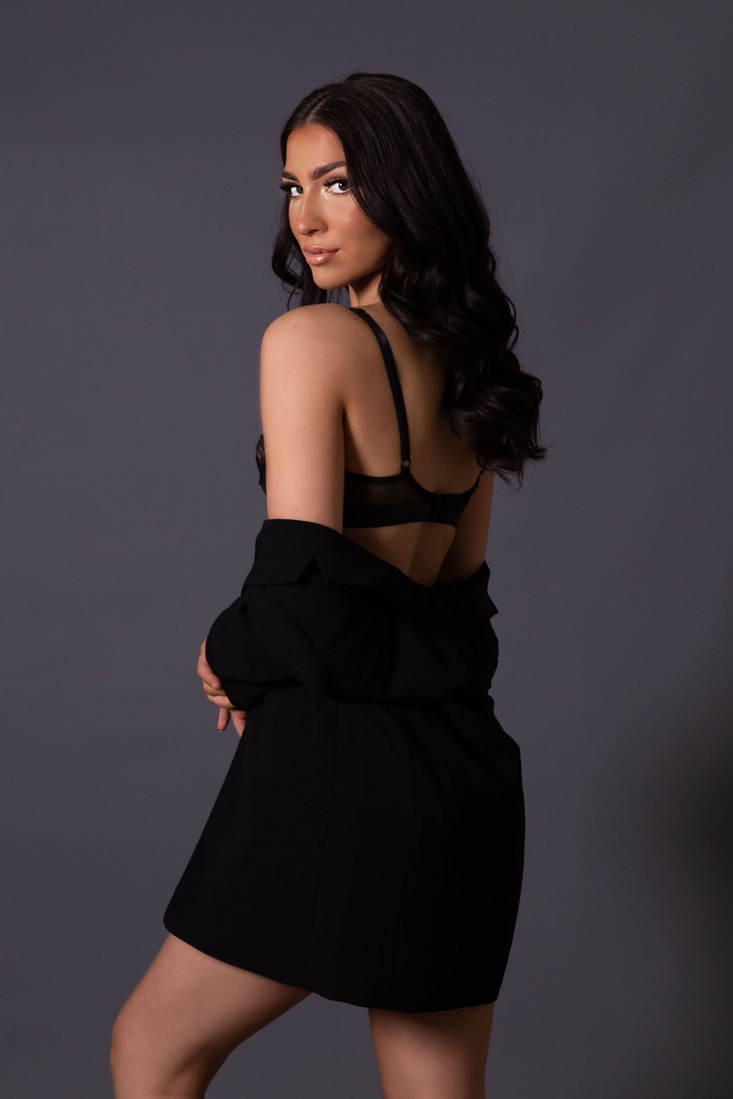 Model in black bra and oversized jacket poses against dark background in glamorous fashion portrait.