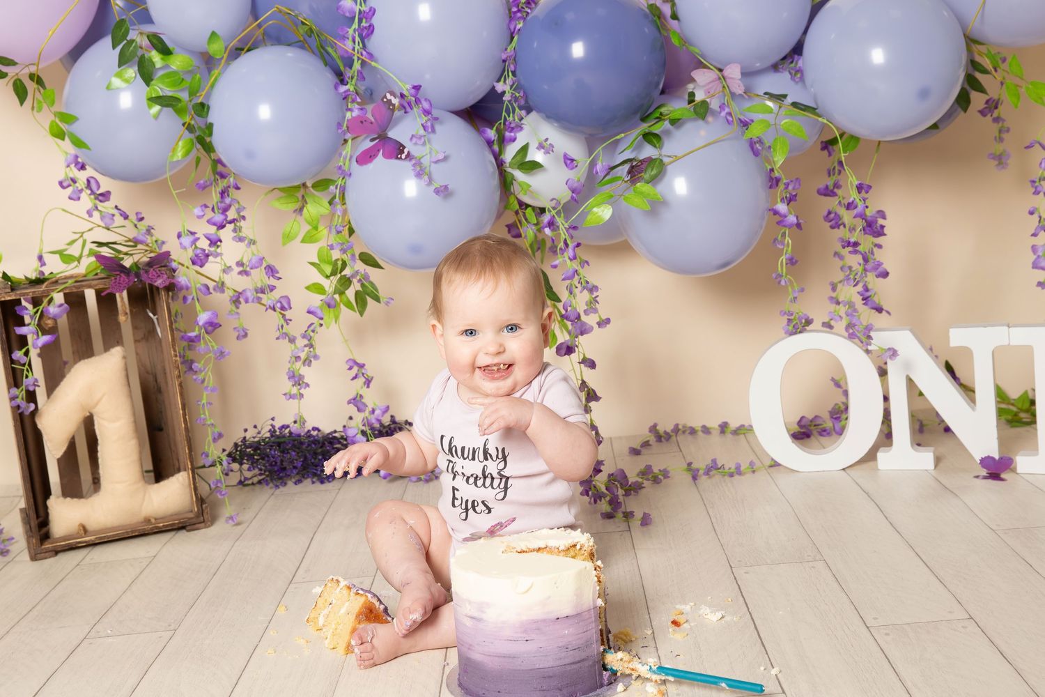 First birthday celebration setup with purple balloons, confetti decorations, and a smash cake on wooden floor.