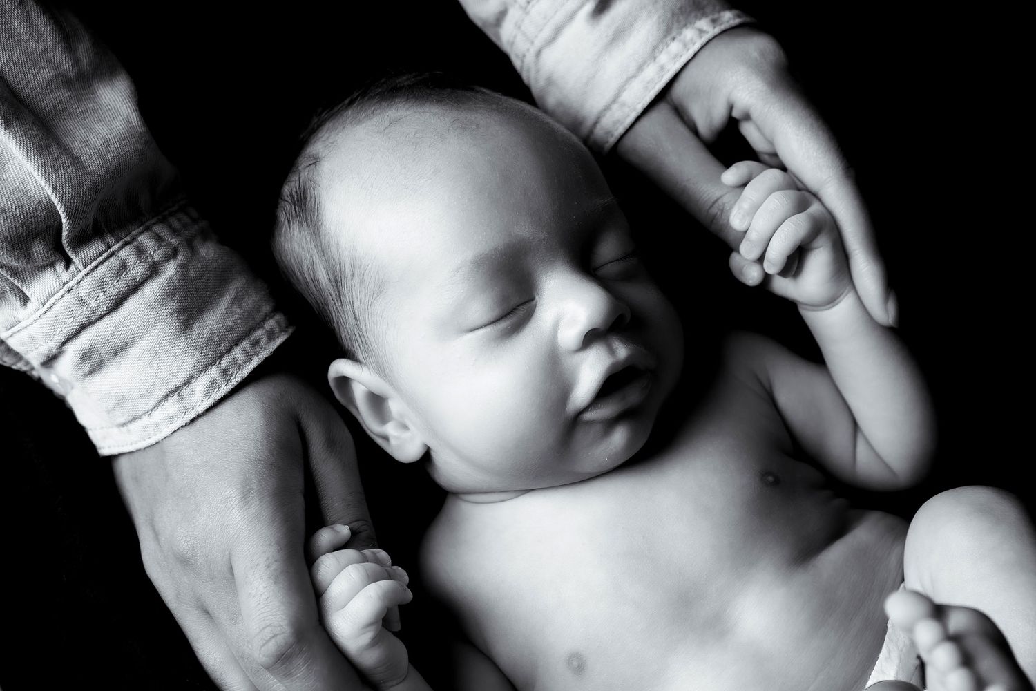 A sleeping infant lies peacefully while grasping adult hands in black and white photography.