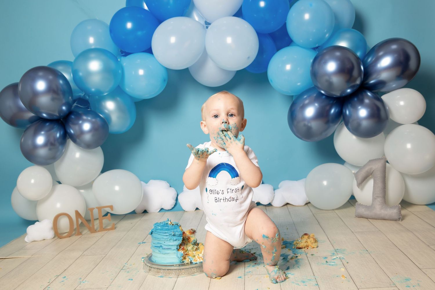 Baby in white outfit sits among blue and silver balloon arch for first birthday cake smash photoshoot.