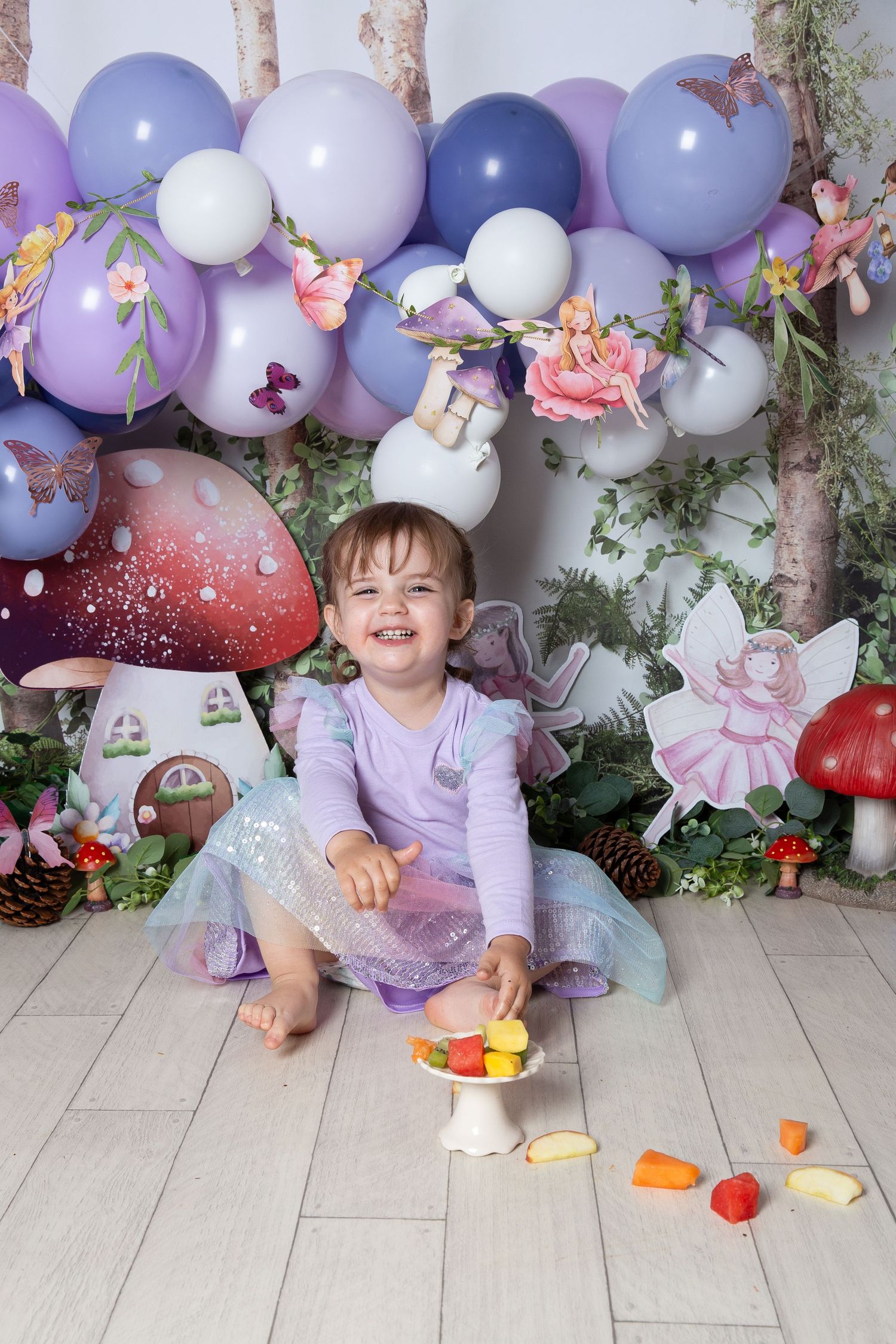 Fairy-themed birthday photo setup with purple balloons, mushroom decorations, and toy fruits scattered on gray flooring.