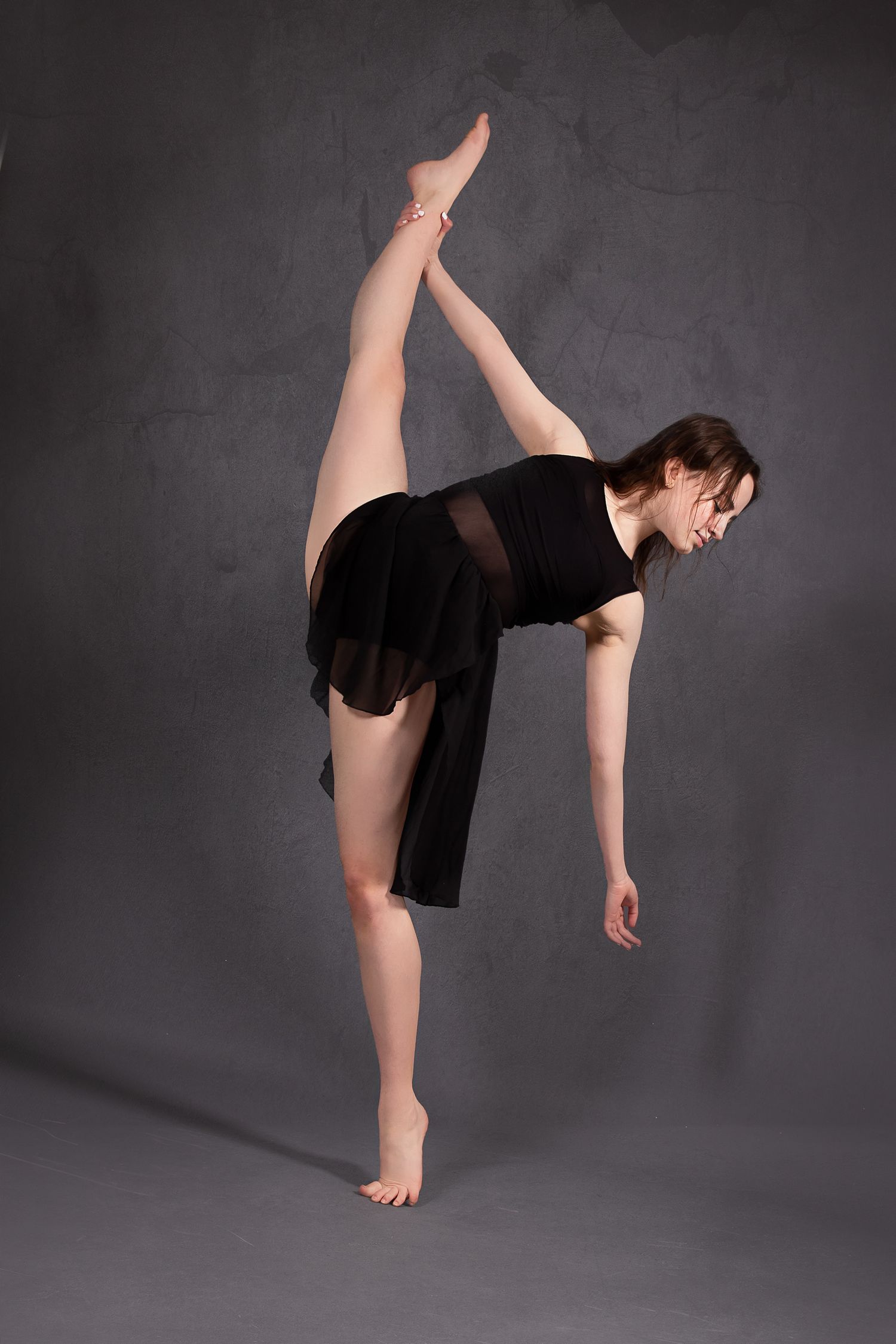 A dancer in a black dress performs a high leg extension against a dark gray background.