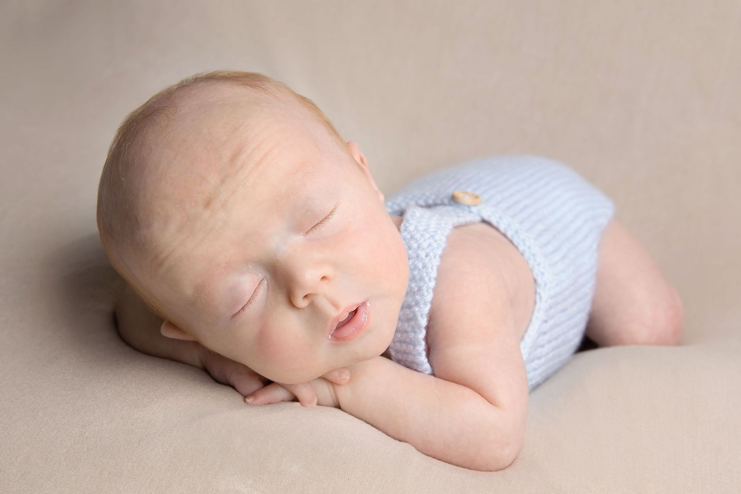 Sleeping newborn wearing light blue romper lying on beige backdrop during professional photo session.