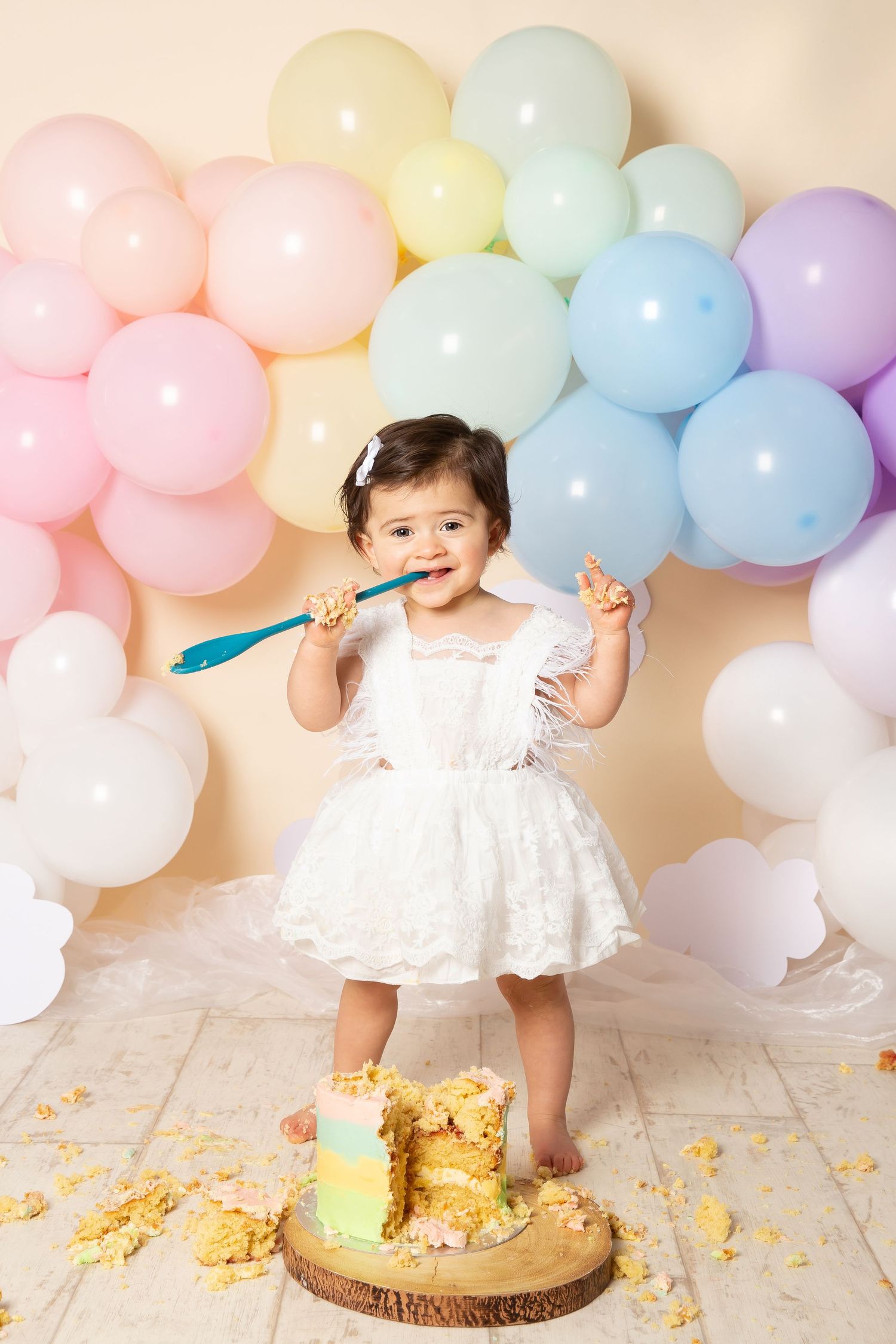 Festive cake smash photo session featuring white dress and pastel balloon garland backdrop with gold confetti accents.