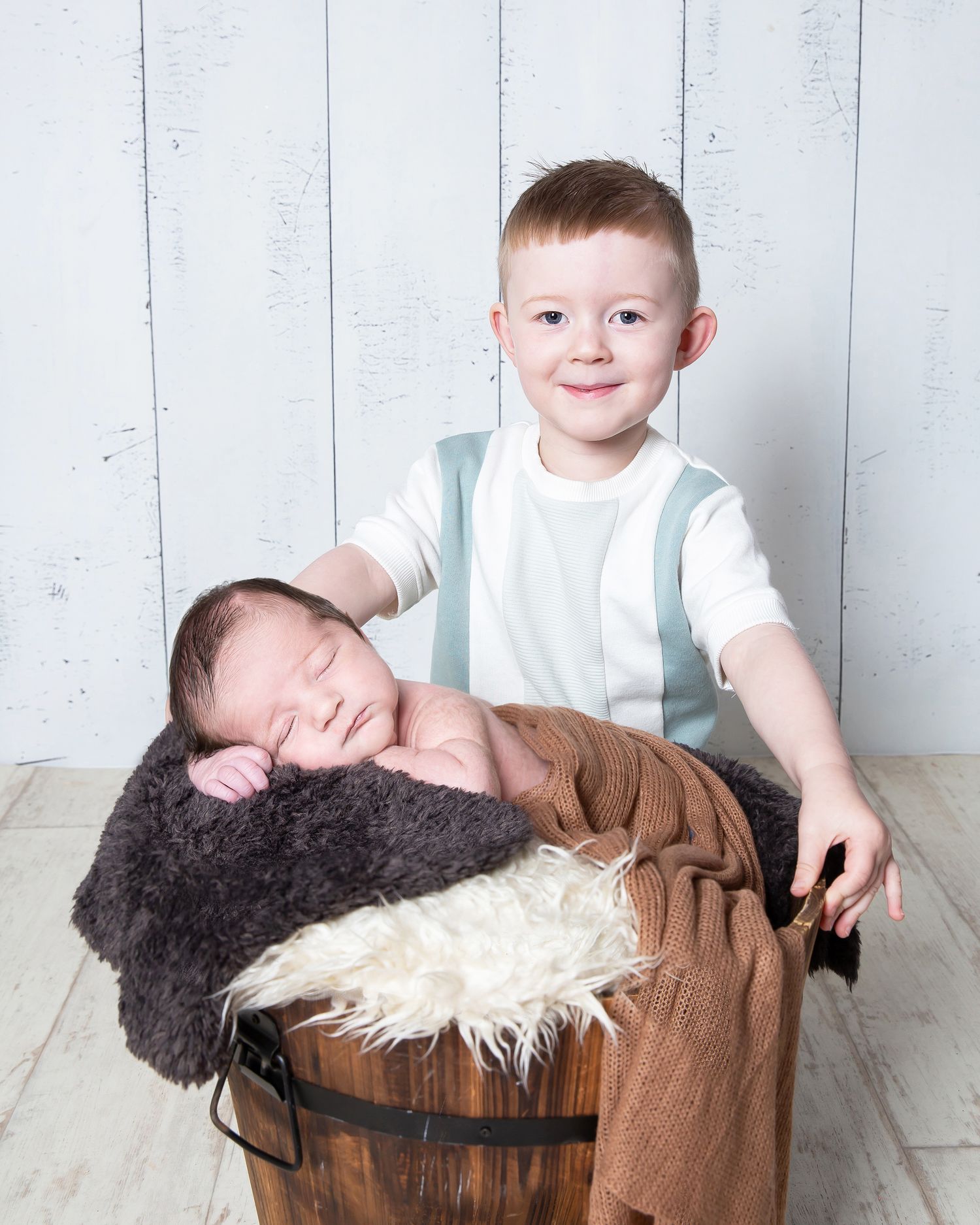 Two children posing for a portrait with rustic barrel and fur prop against white wooden background.