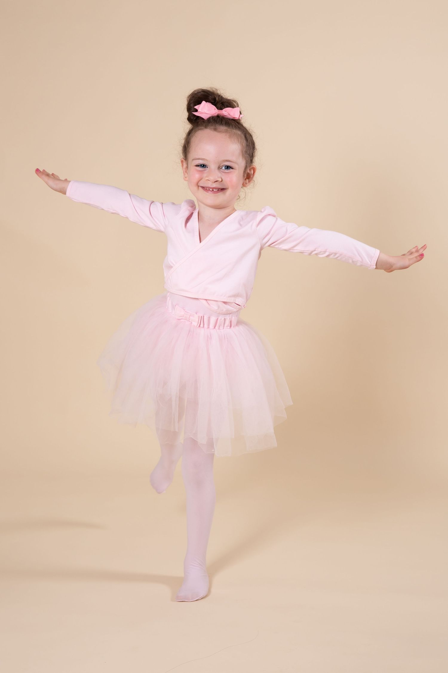 Small dancer in pink wrap top and tulle skirt strikes joyful pose with arms outstretched.
