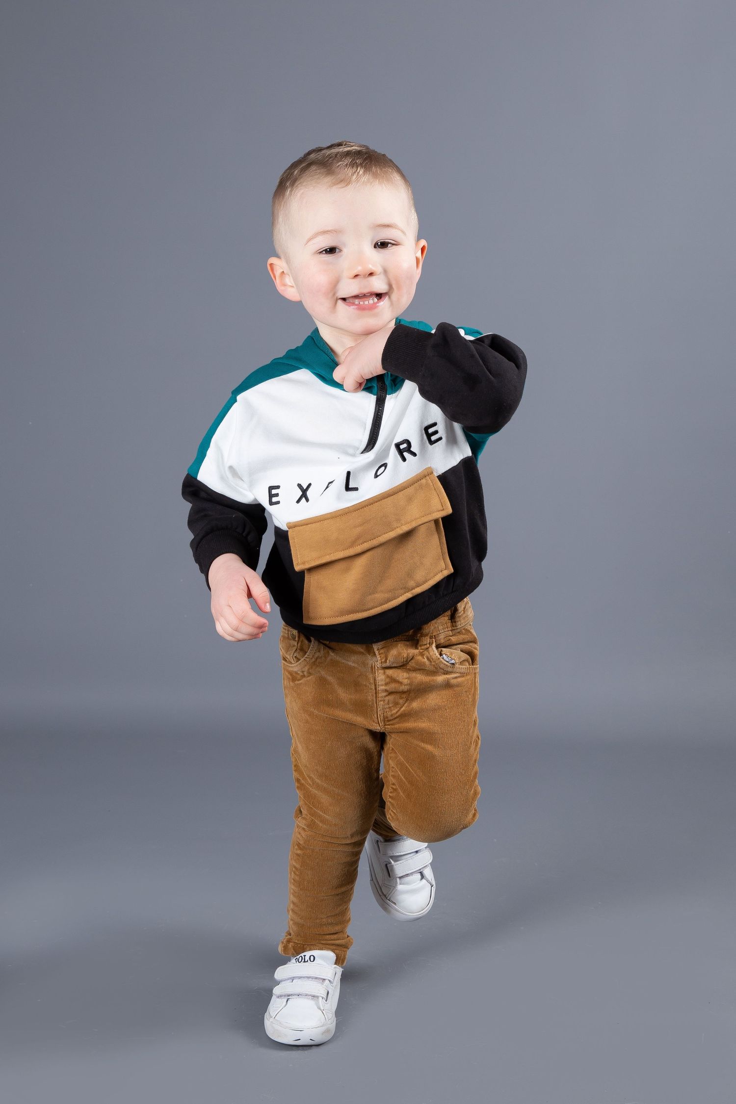 A cheerful toddler wearing a color-block jacket and brown pants strikes a fun pose against a gray background.
