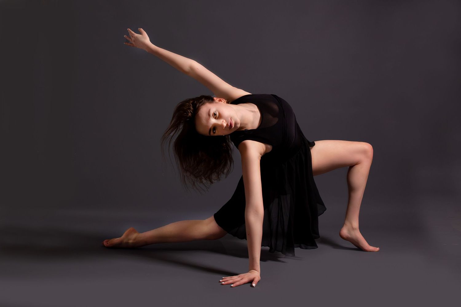 Graceful dancer in black dress performs modern dance movement on dark studio floor.