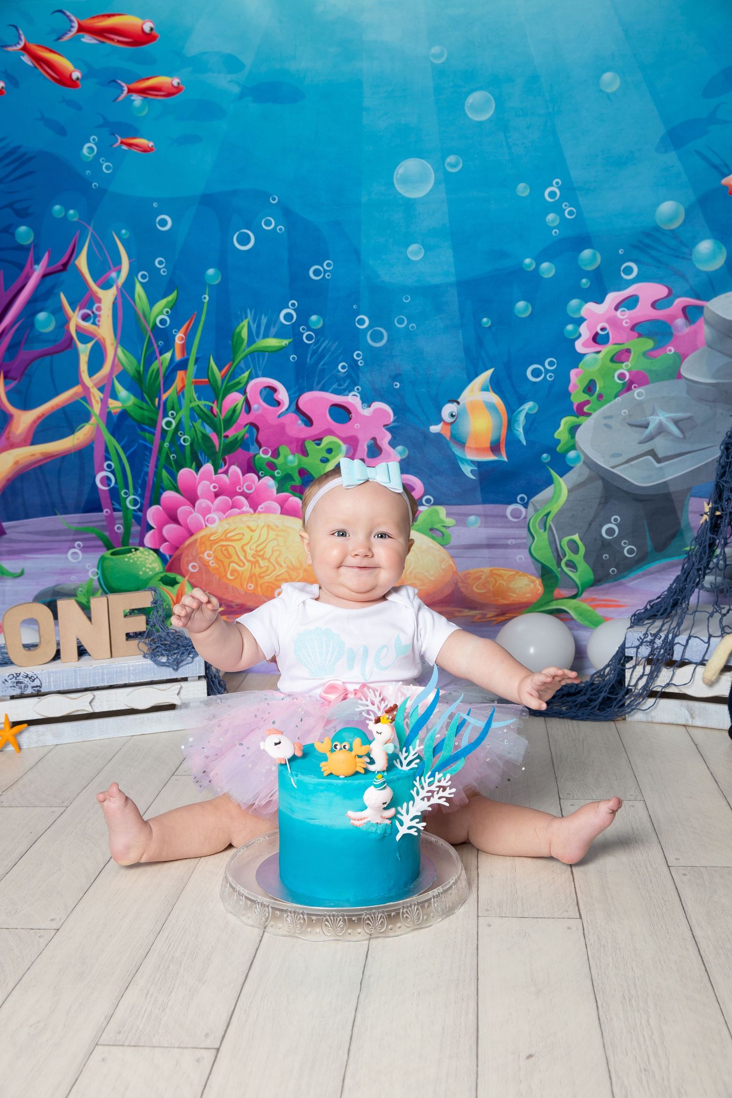 Underwater themed cake smash photo session setup with blue backdrop, coral decorations, and bubbles for baby milestone photos.