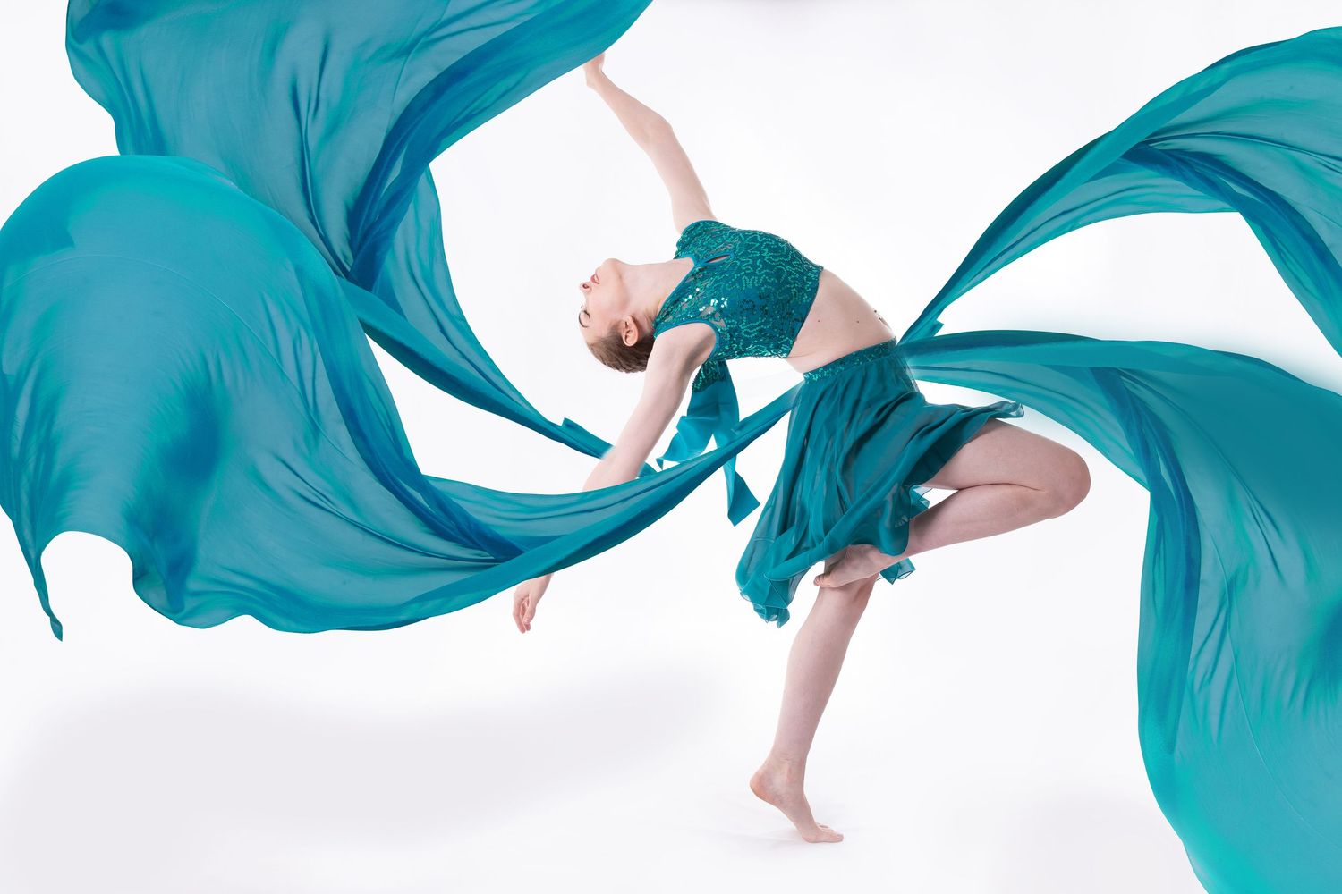 Dancer in flowing turquoise fabric creates graceful movement against white background in artistic performance shot.