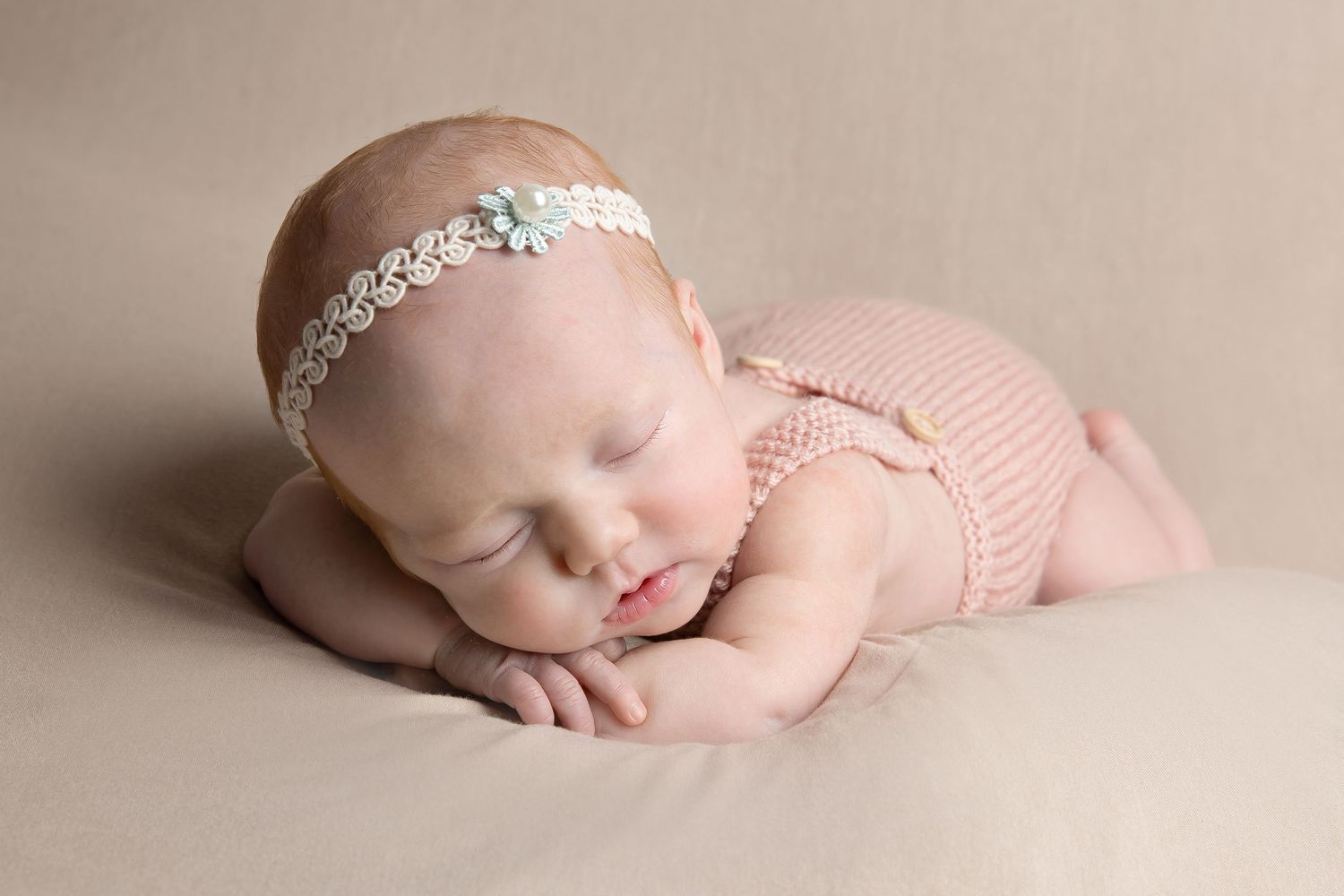 Peaceful sleeping infant in pink outfit with lace headband resting on neutral backdrop.