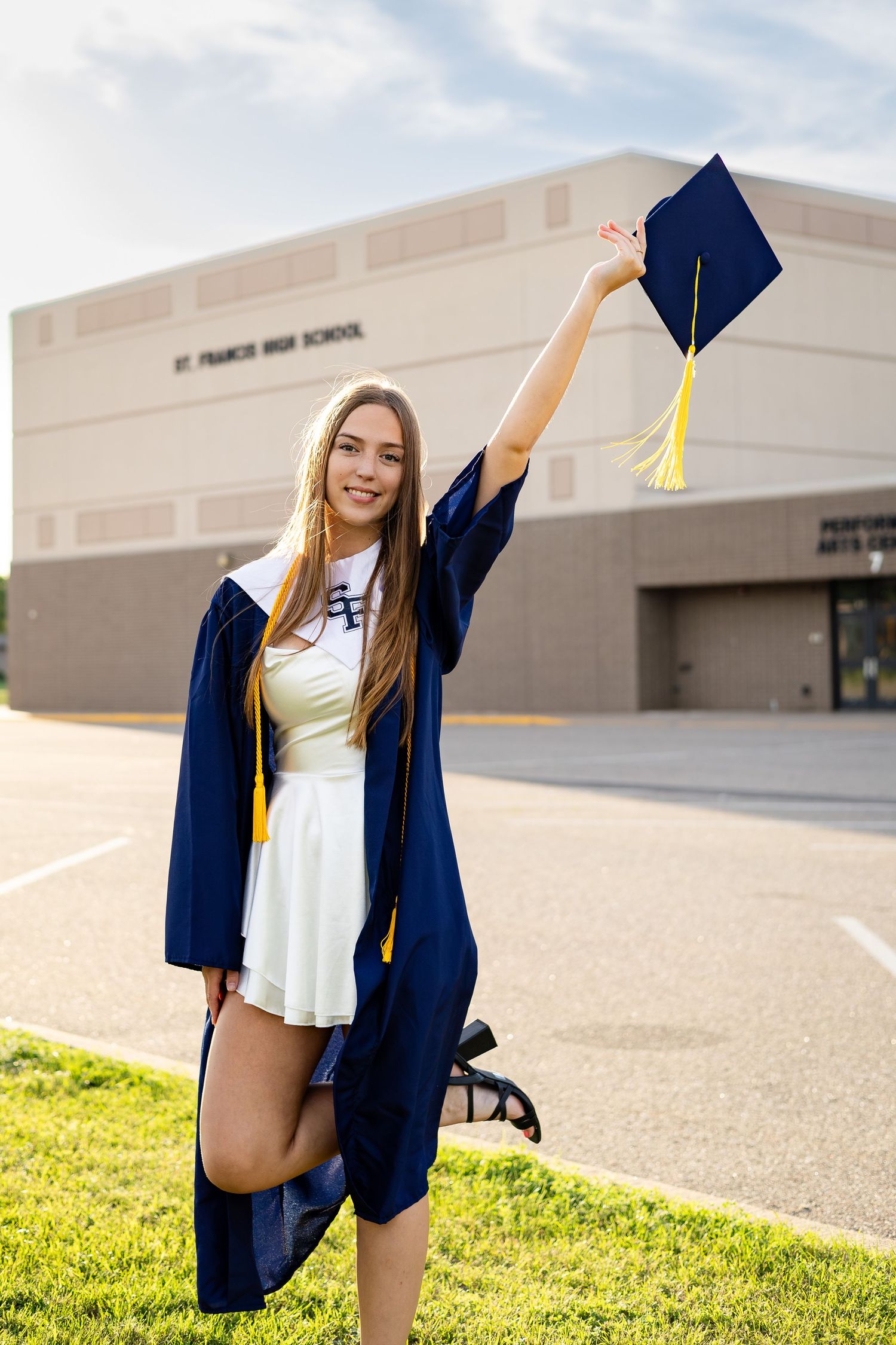 Graduate posing with cap and gown against school building exterior in sunlit graduation photoshoot.