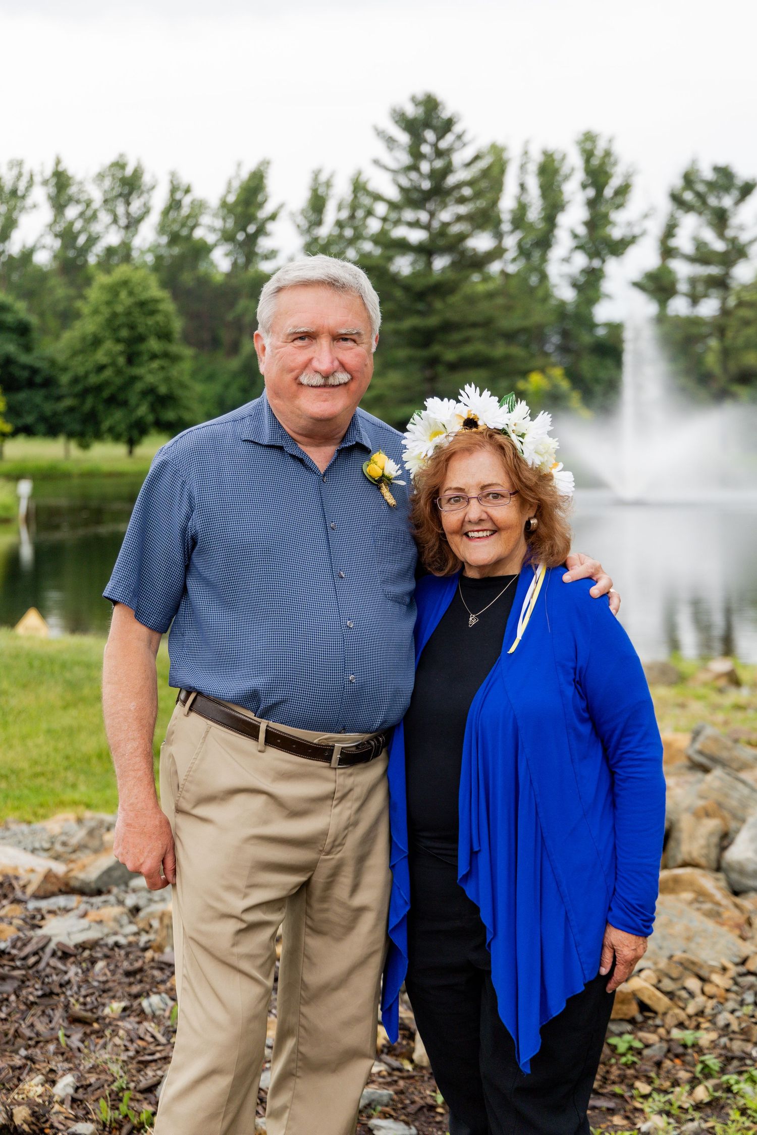 A couple poses outdoors by a fountain wearing coordinated blue and khaki outfits with floral accessories.