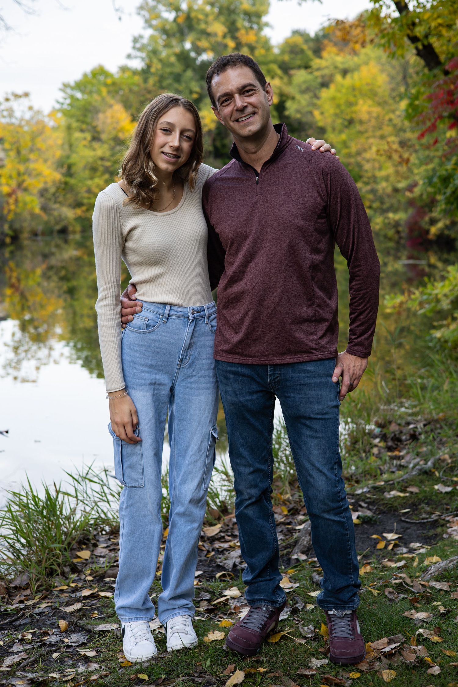 Couple standing together on leaf-covered shore with lake in background.