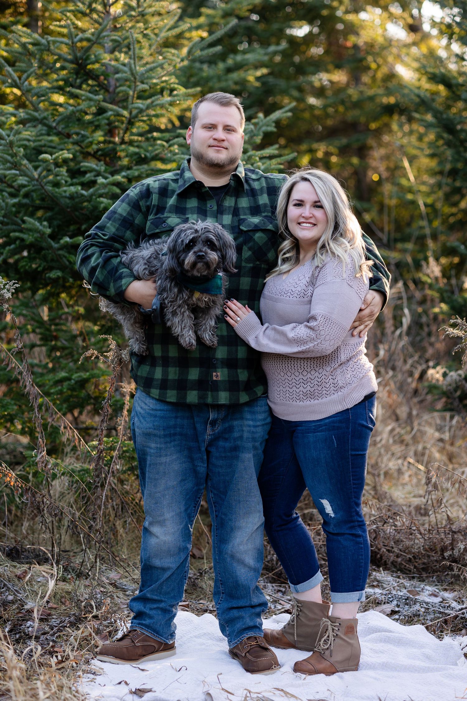 A couple in winter attire posing together on snowy ground with evergreen trees in the background.