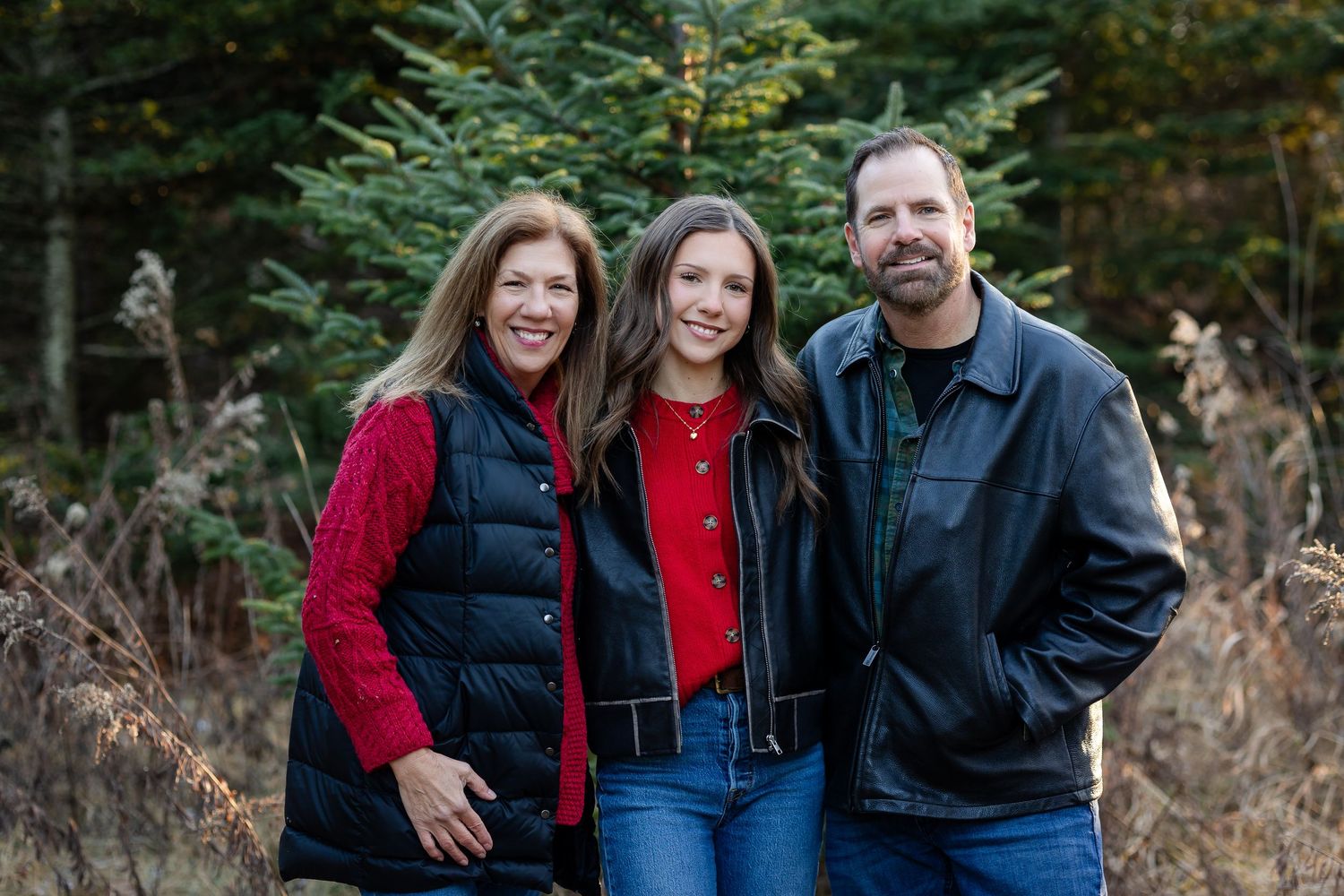 Family wearing red and black clothing poses together outdoors in a wooded winter setting.