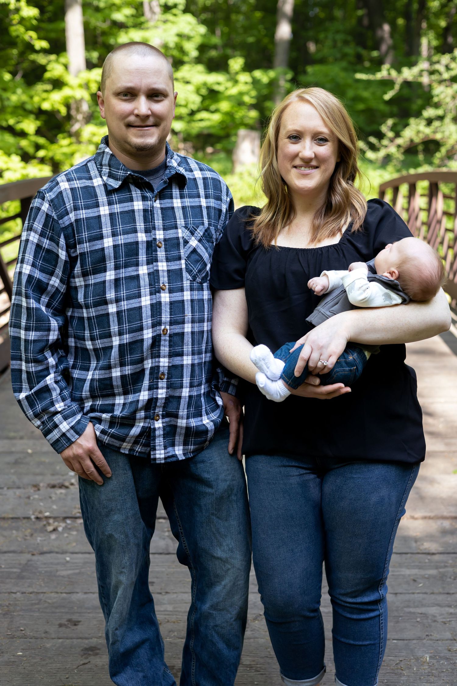 Family portrait on wooden bridge with parents in casual attire holding infant in wooded setting.