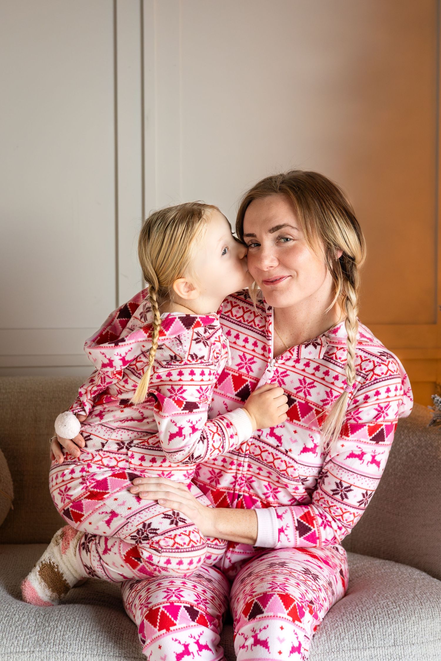 Mother and child in matching pink Nordic print Christmas pajamas sharing tender moment on sofa.