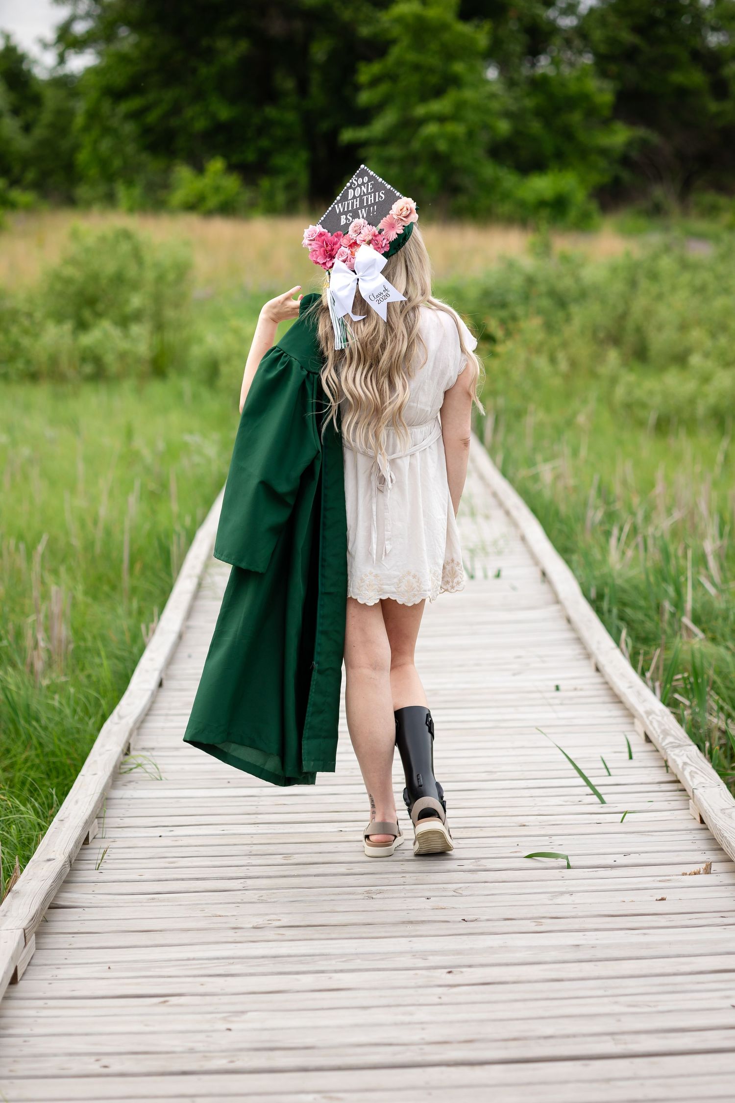 Blonde graduate walking wooden boardwalk path wearing decorated cap and carrying green gown.
