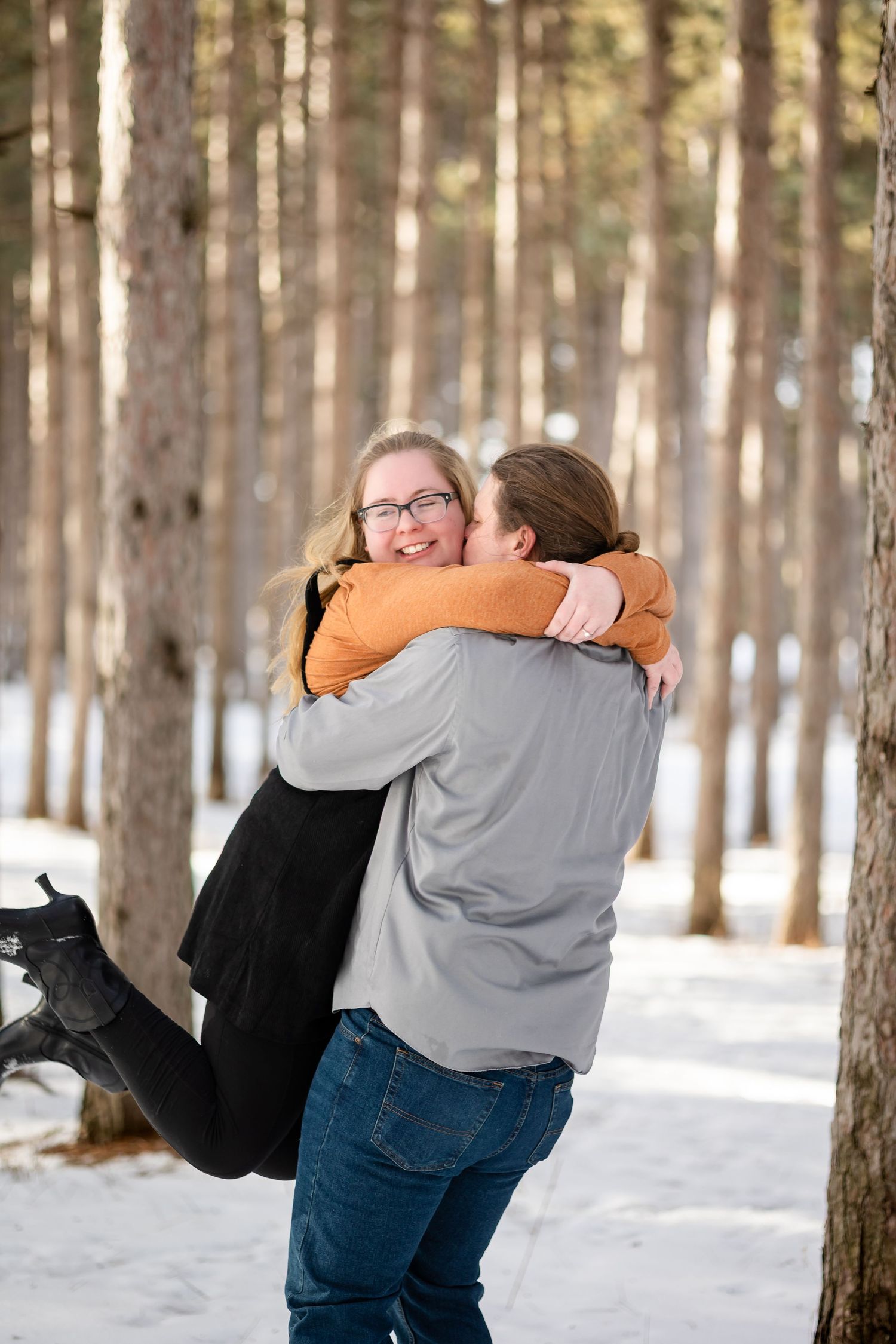 Couple hugging joyfully in a snowy pine forest, woman smiling with glasses, man in gray shirt and jeans.