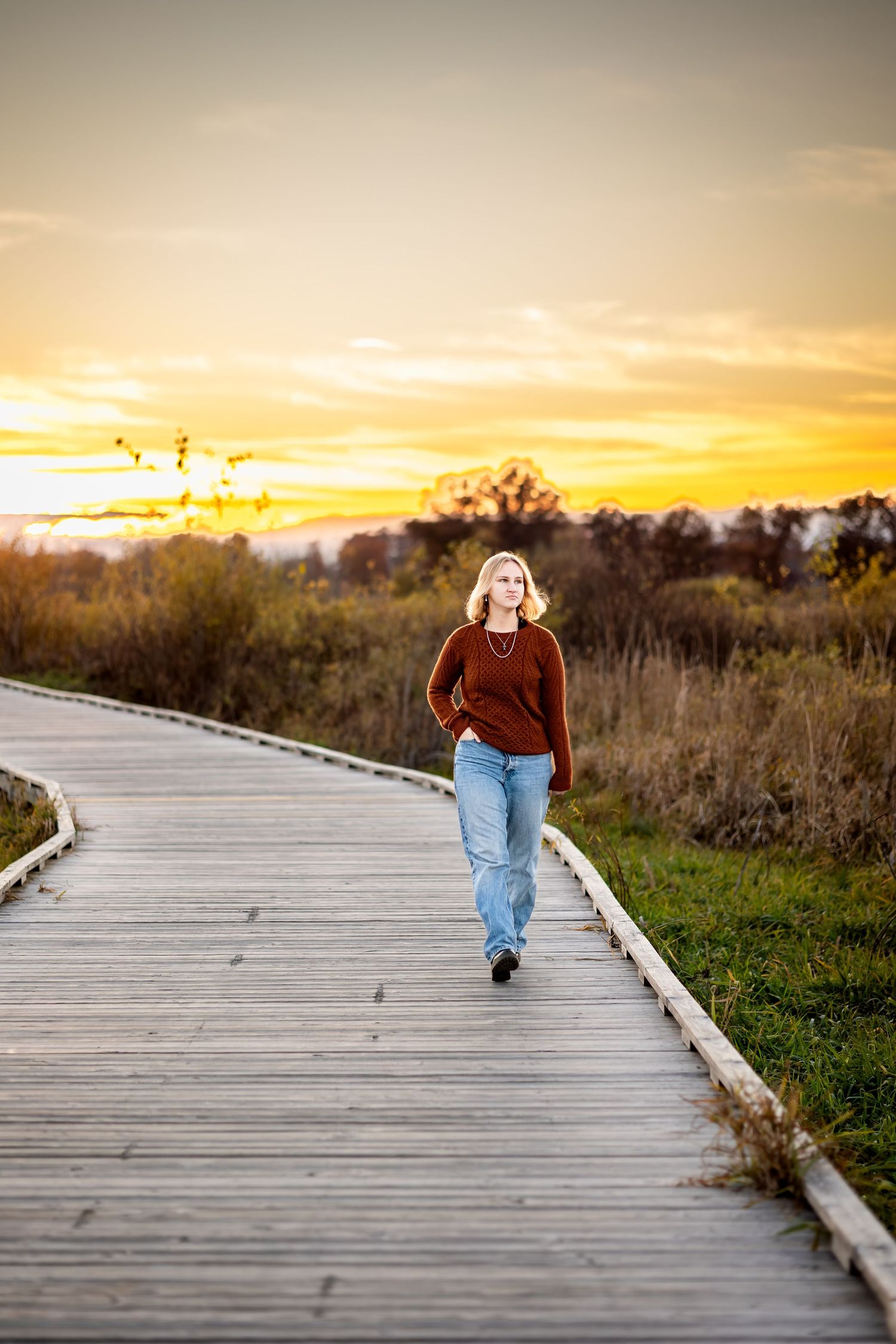 Person in jeans walking away on a wooden boardwalk at sunset with golden sky and trees in background.