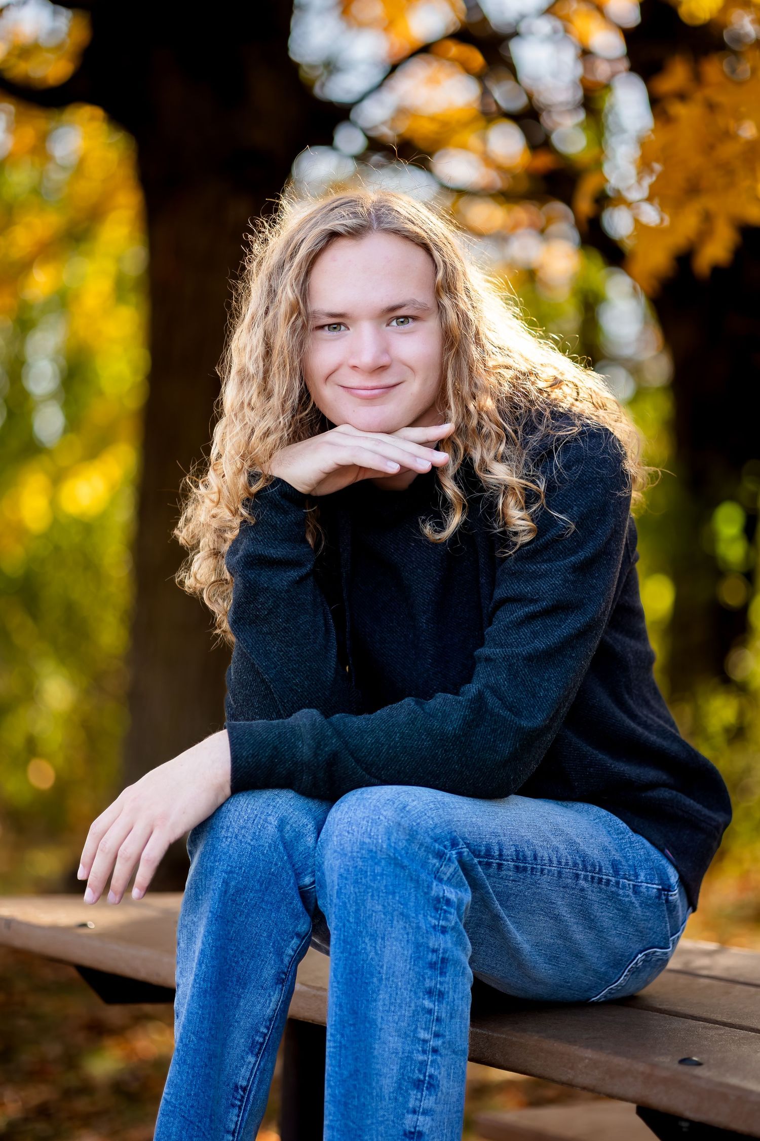A casual portrait in autumn with someone wearing a black sweater and jeans sitting on a wooden bench surrounded by fall foliage.