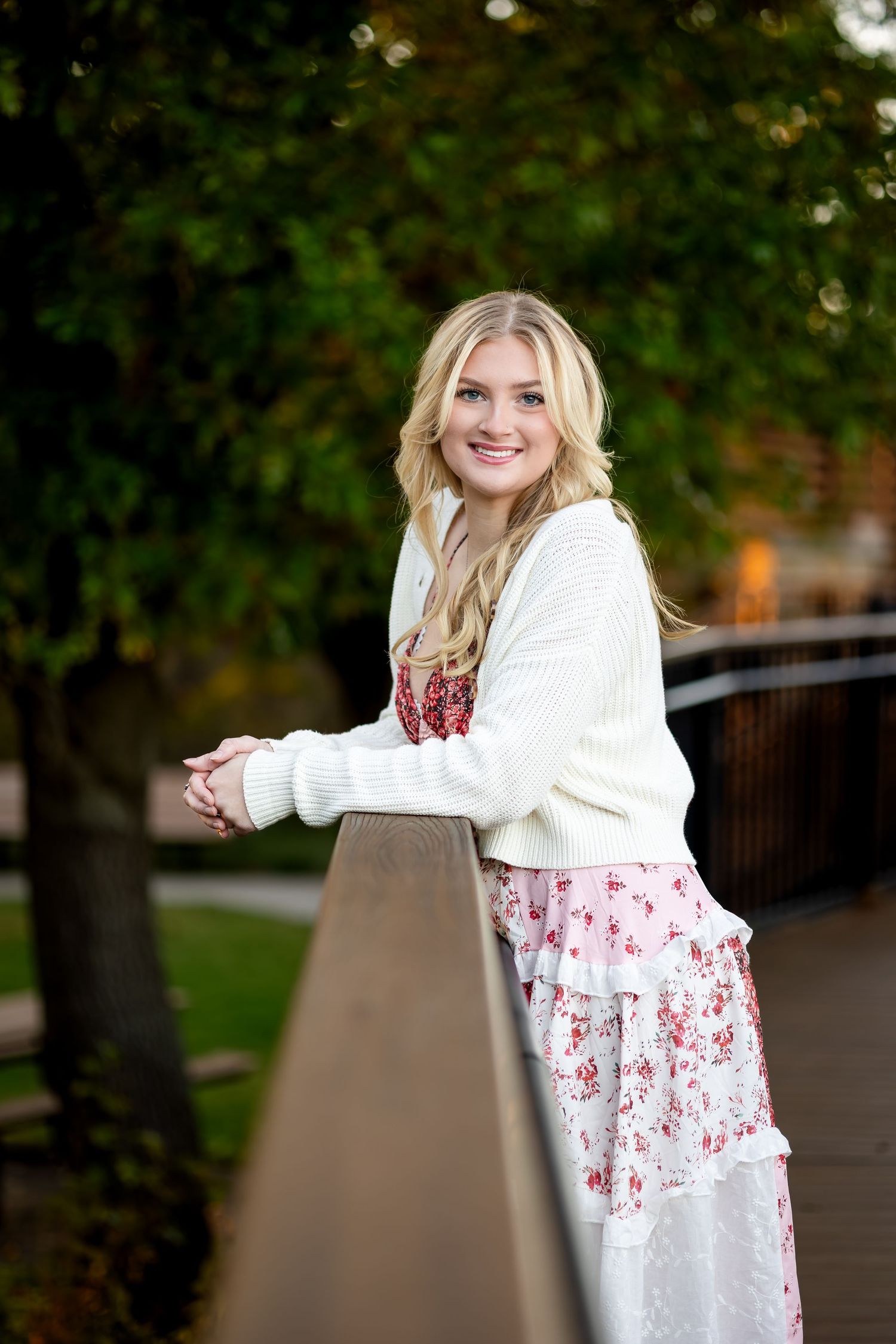 A person in a white sweater and floral dress leans against a railing in an outdoor park setting.