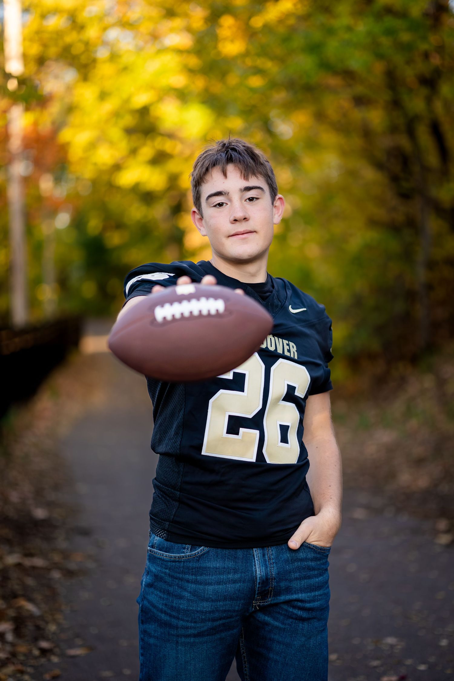 A football player in a black jersey with number 26 poses with a football against an autumn backdrop with yellow foliage.