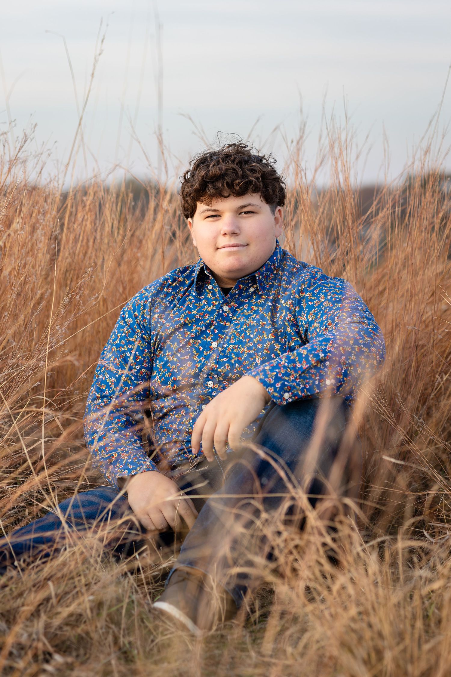 Person in blue patterned shirt sitting among dried autumn grass.