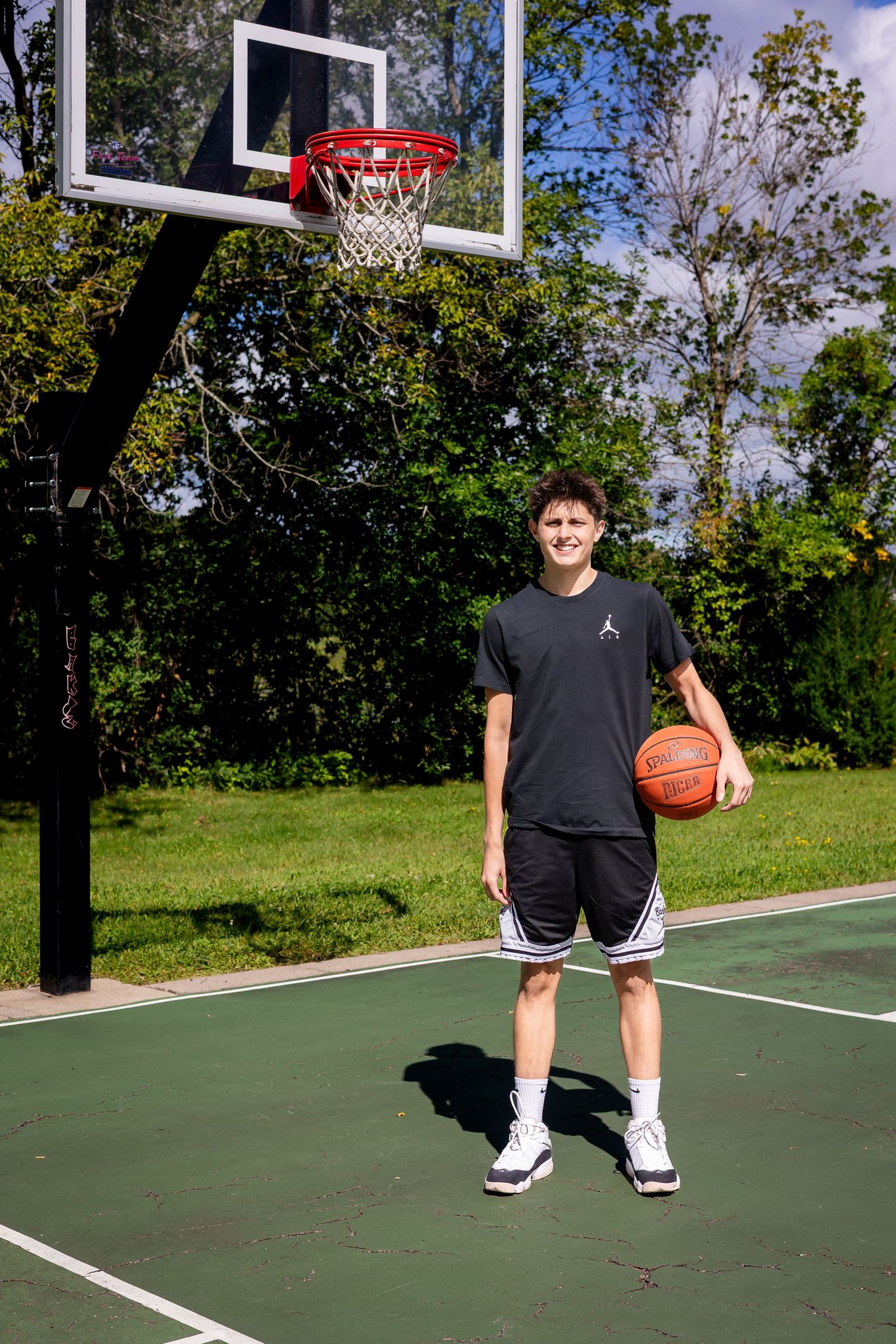 Basketball player on outdoor court with hoop and green surroundings.