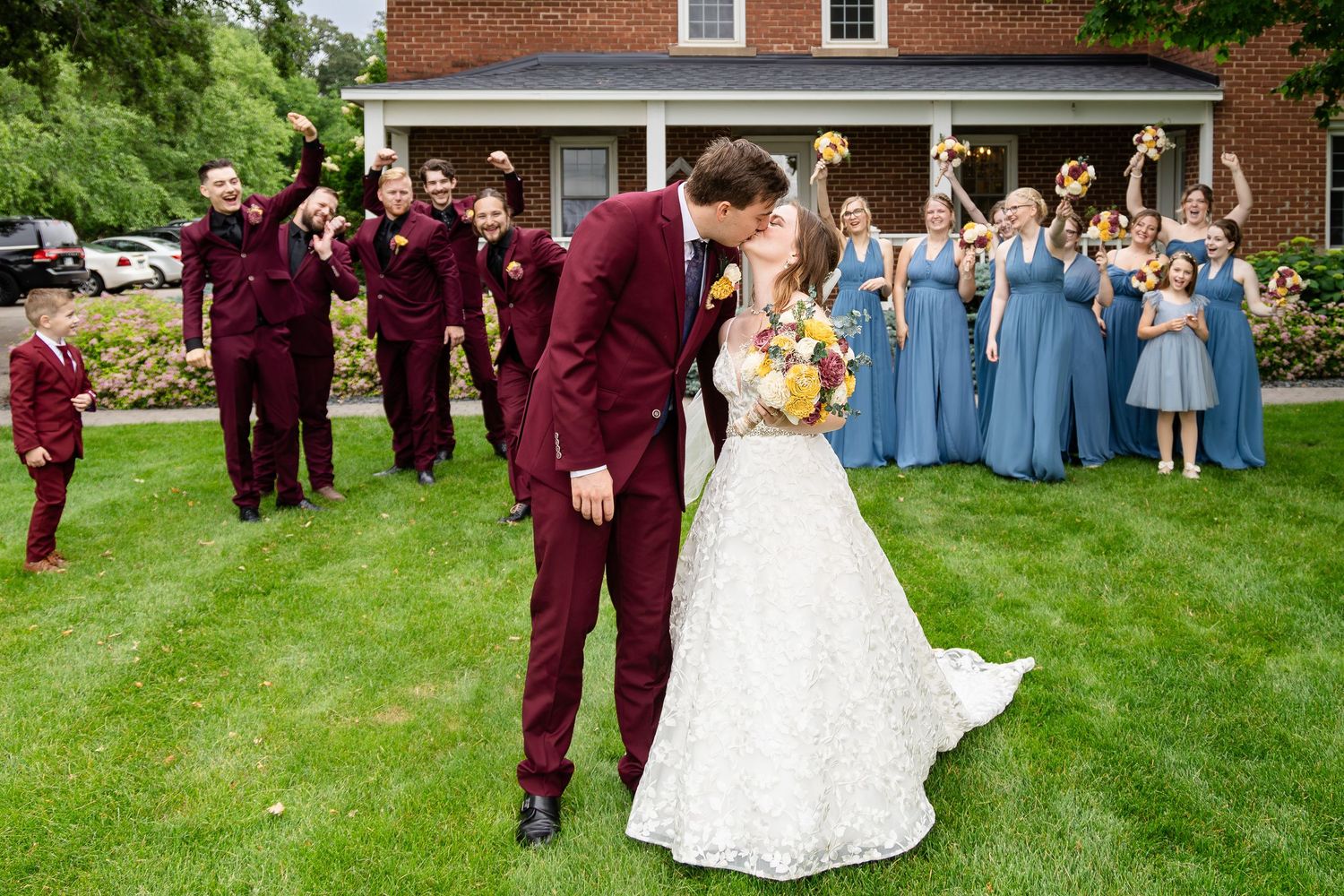 Wedding party poses on green lawn in front of brick building with burgundy and blue attire.