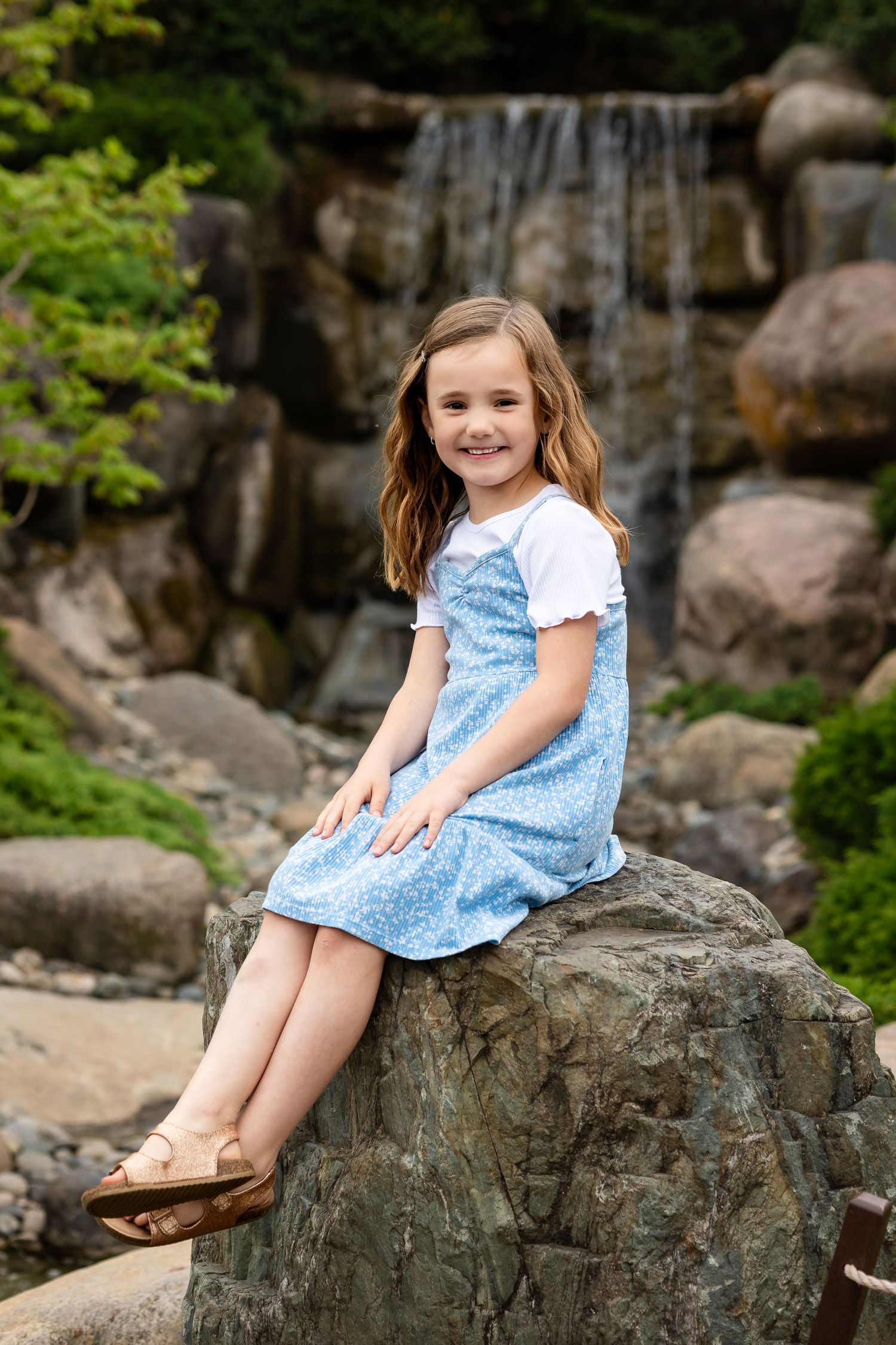 Smiling young girl in a blue floral dress sits on a large rock with a scenic waterfall backdrop.