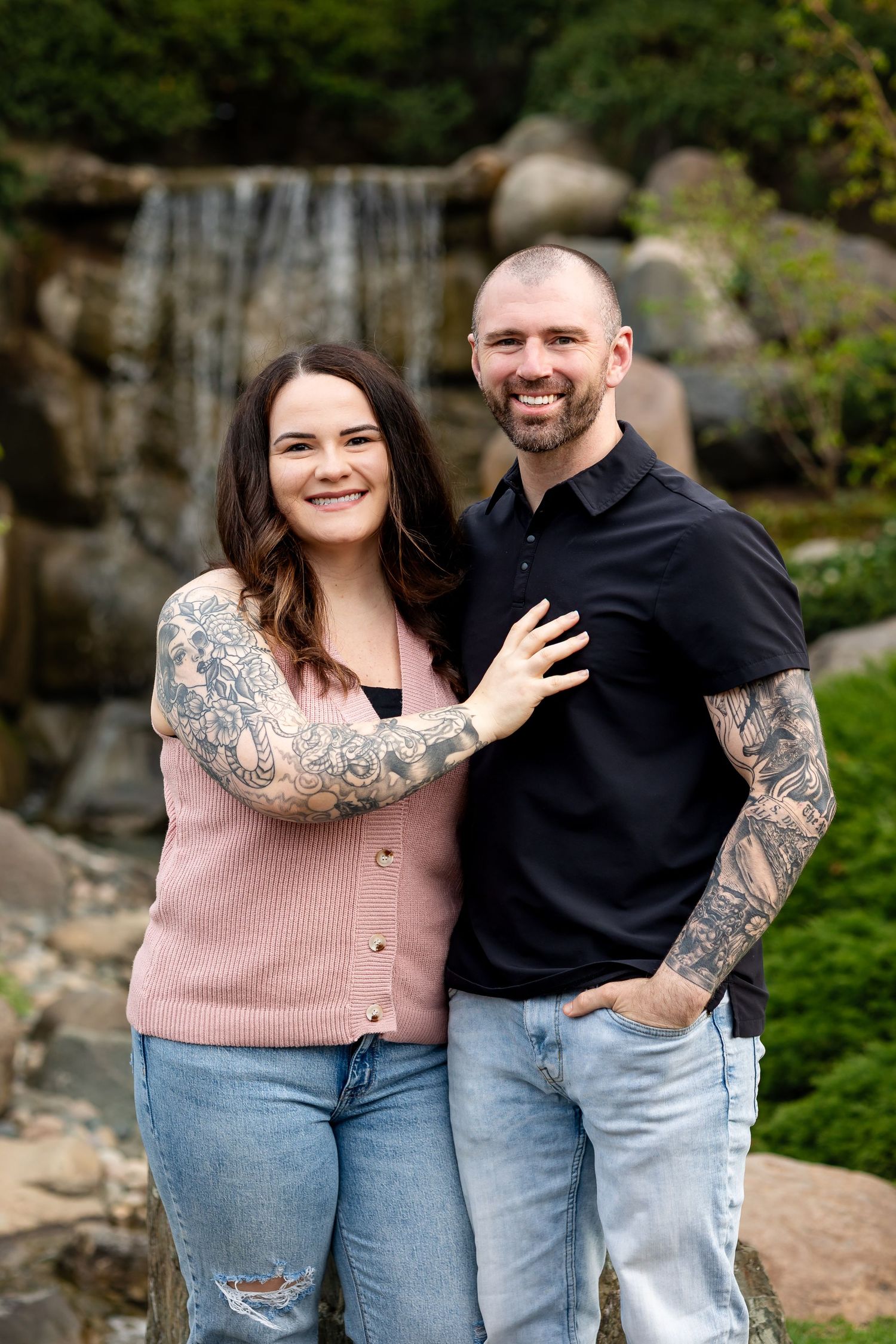 Tattooed couple smiling together in front of a rocky waterfall in a lush garden.