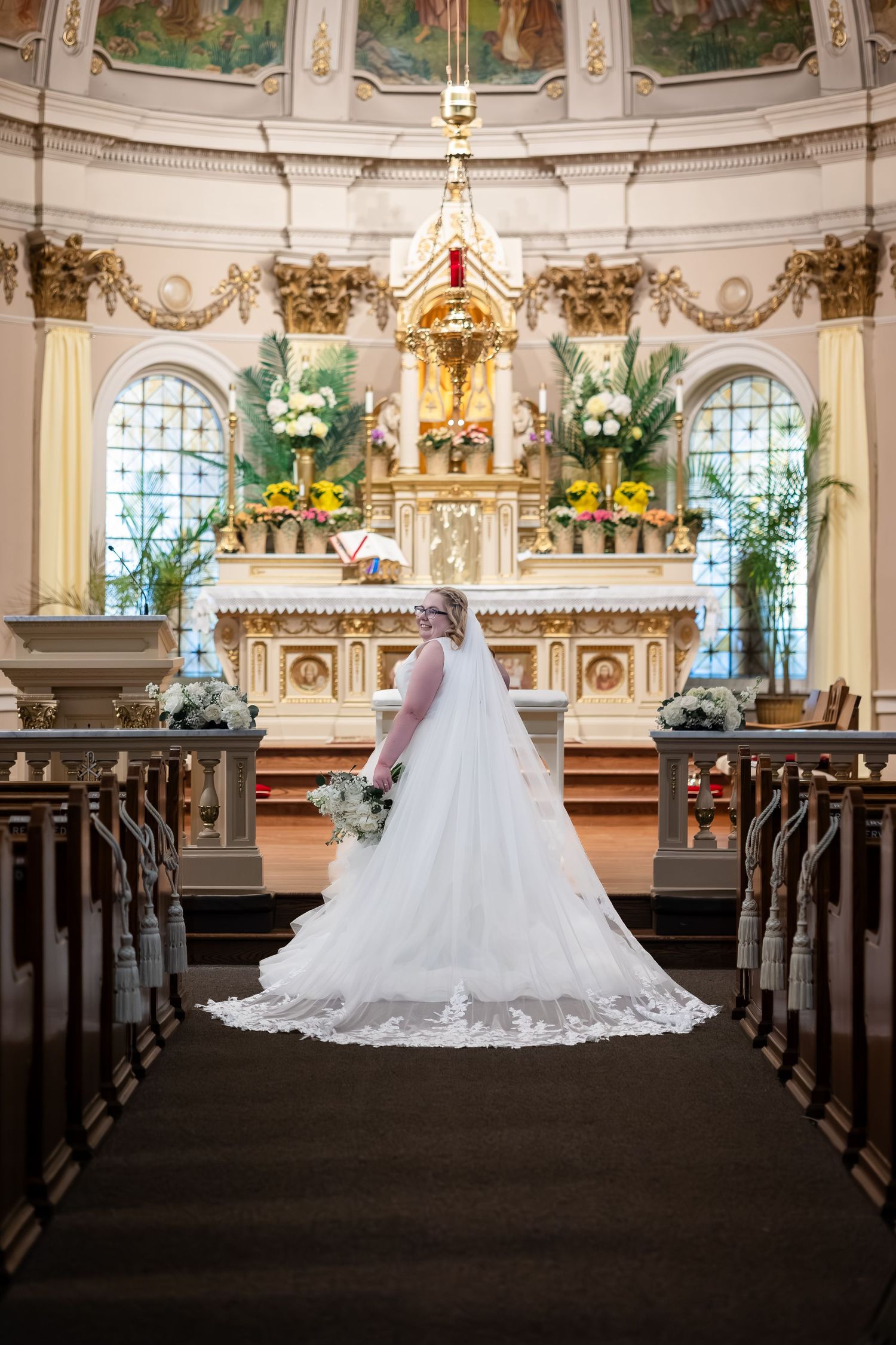Bride in white lace gown with cathedral train stands alone at ornate church altar holding bouquet.