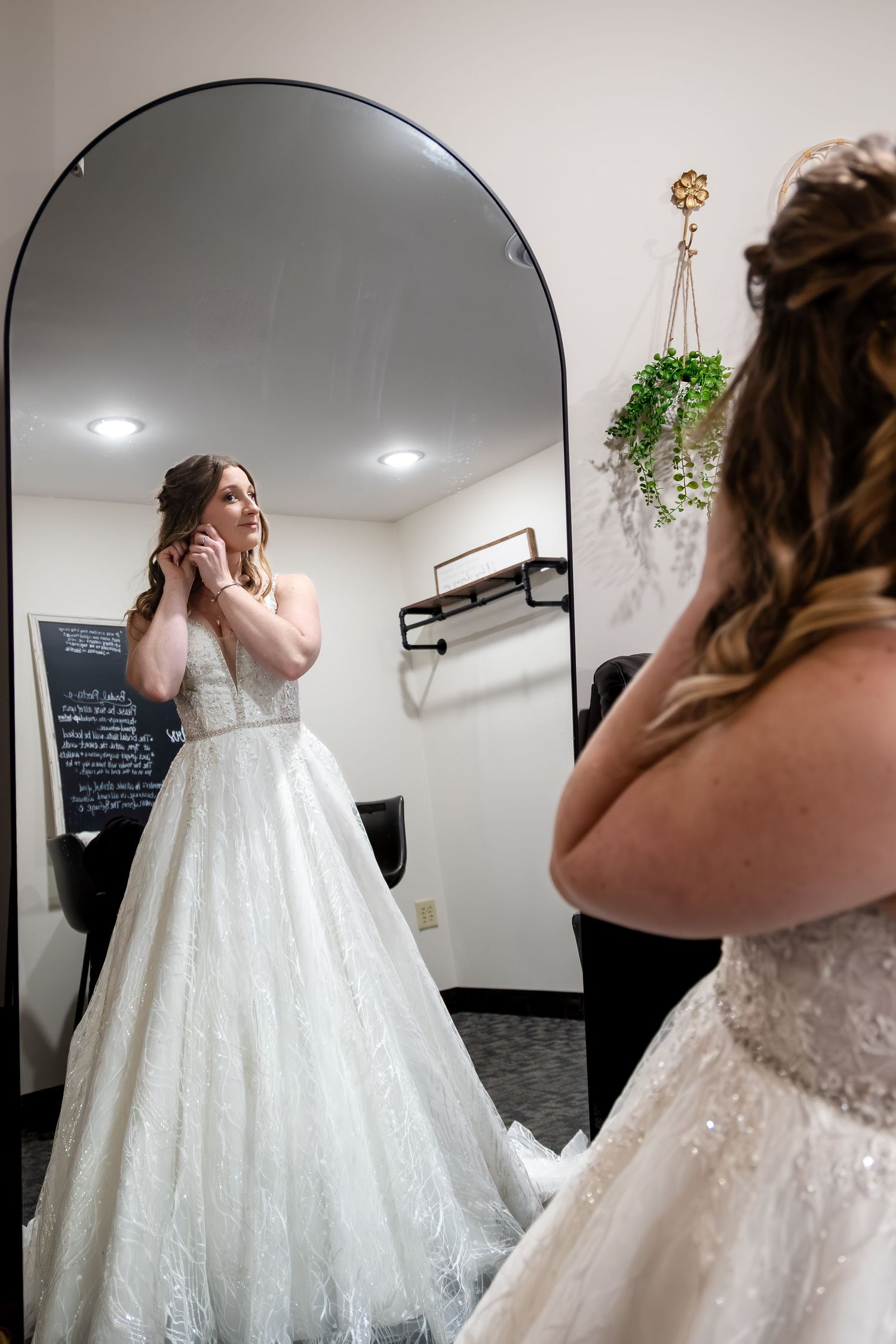 Bride in a sparkling ball gown adjusts her earring while gazing at her reflection in an arched mirror.