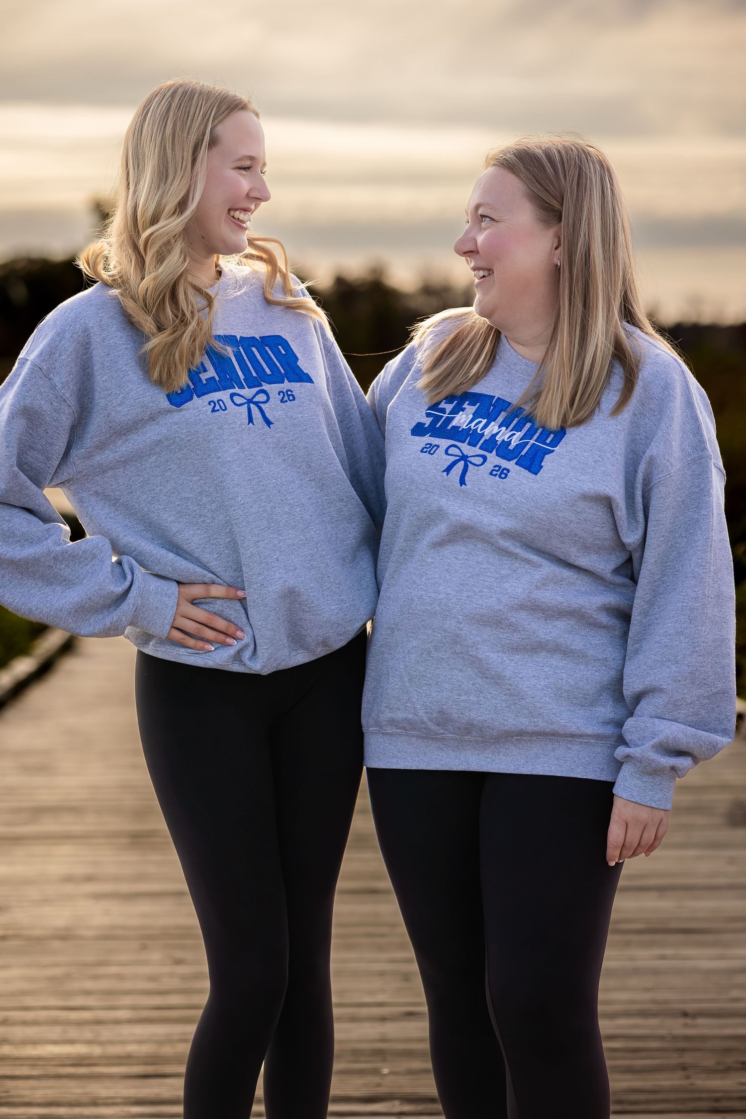 Two people in matching gray sweatshirts share a laugh while standing on a wooden dock at sunset.