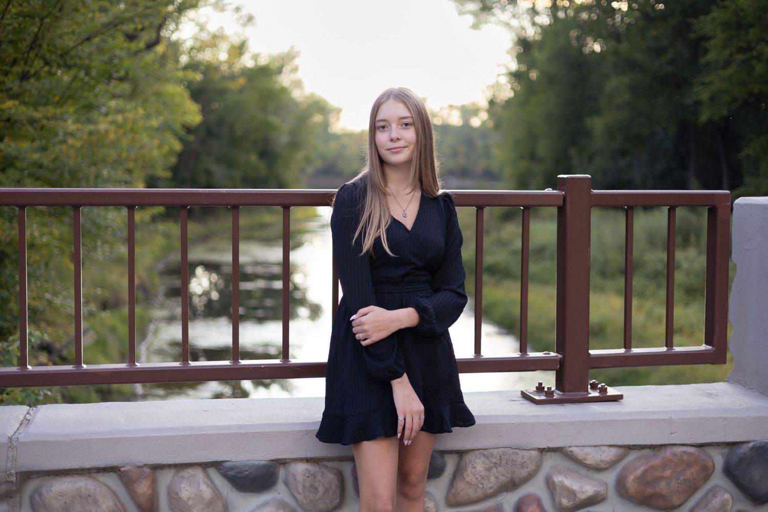 Person in black dress standing near stone wall and railing at sunset.