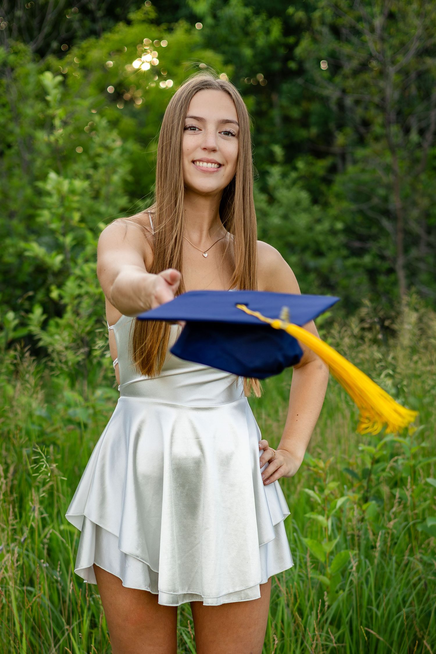 Graduate in white dress poses playfully with blue cap in grassy outdoor setting.