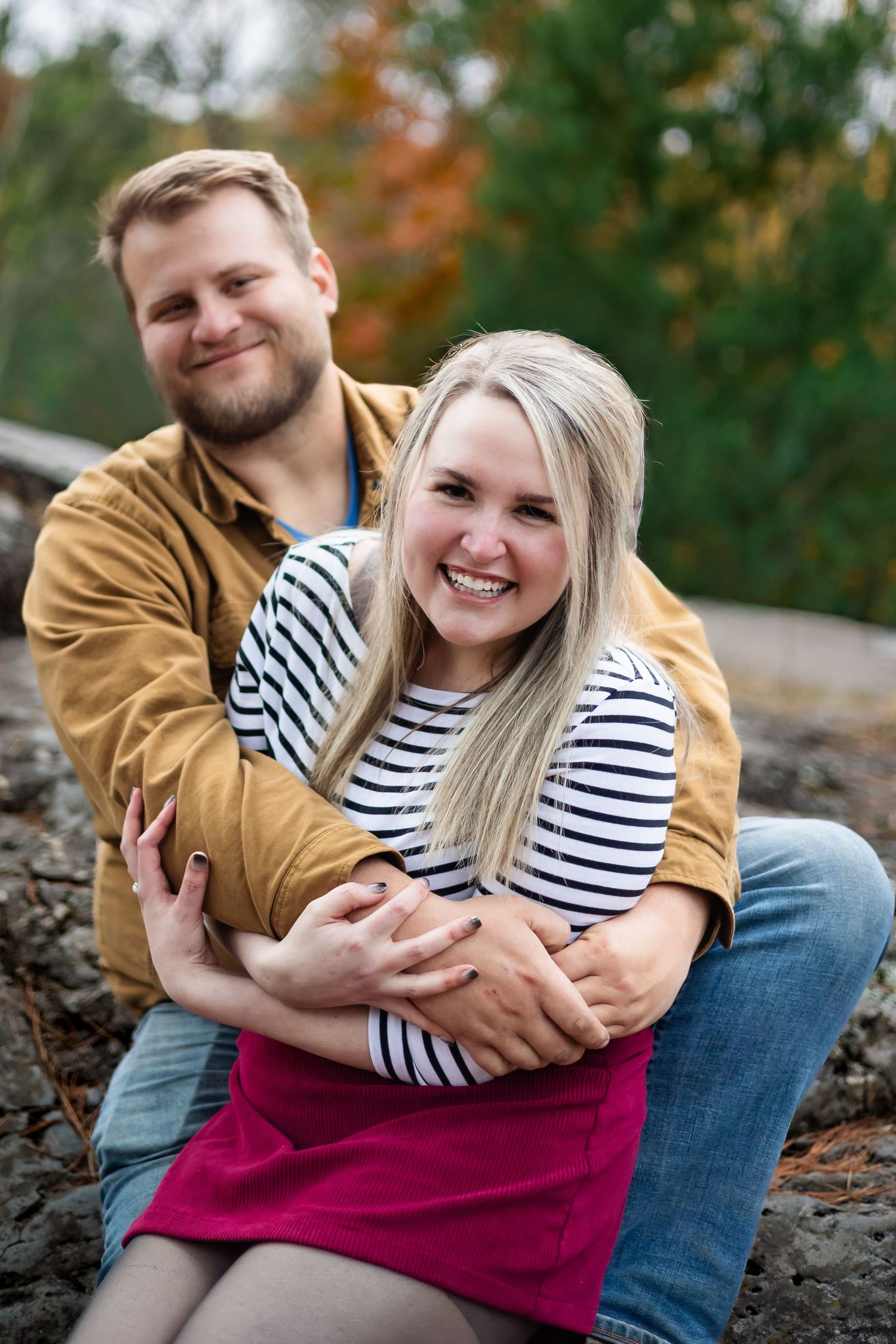 A smiling couple embraces outdoors in autumn, woman in striped shirt and pink skirt, man in tan jacket.
