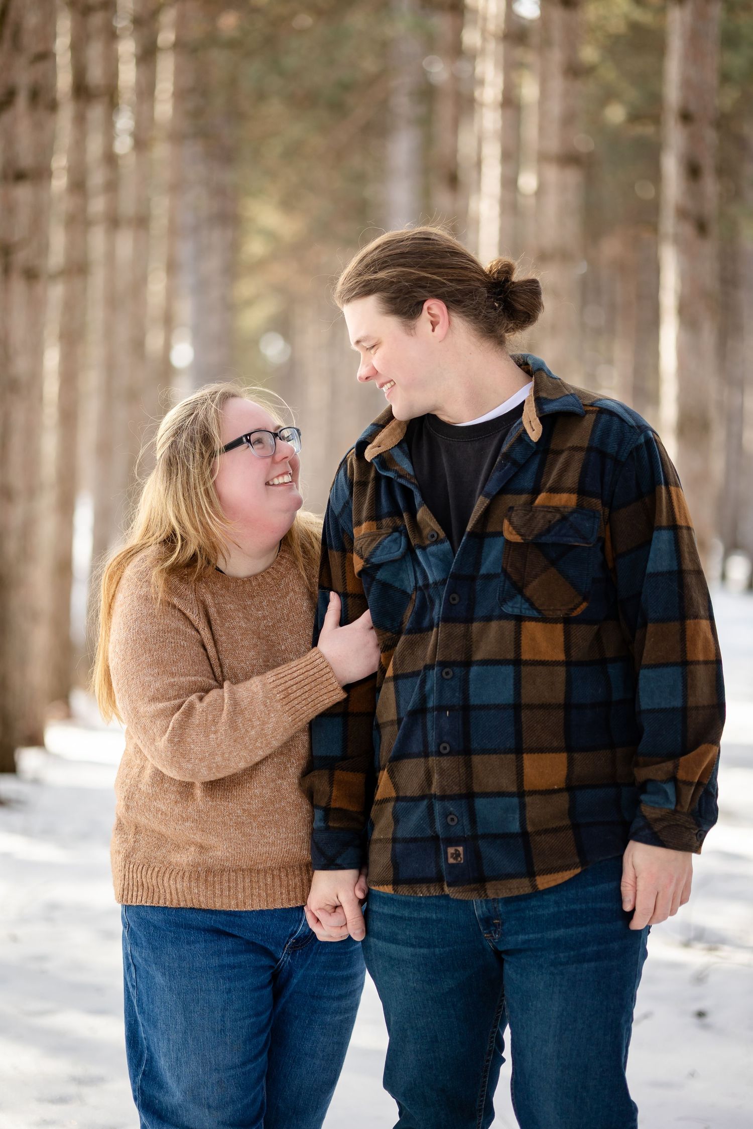 Couple holding hands and smiling at each other in a snowy pine forest, wearing casual winter clothing.