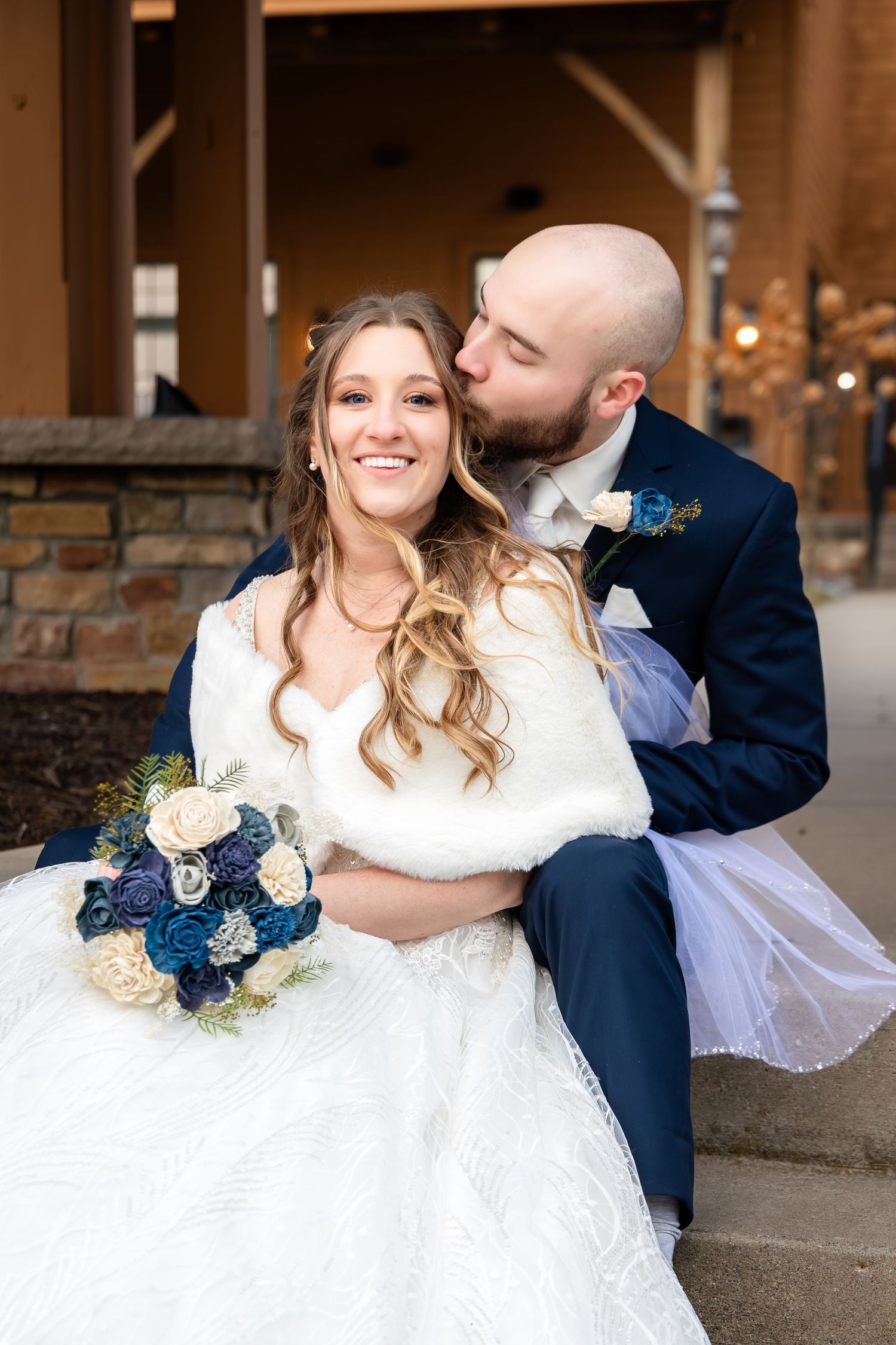 Bride smiling with bouquet as groom kisses her cheek in navy suit outdoors.