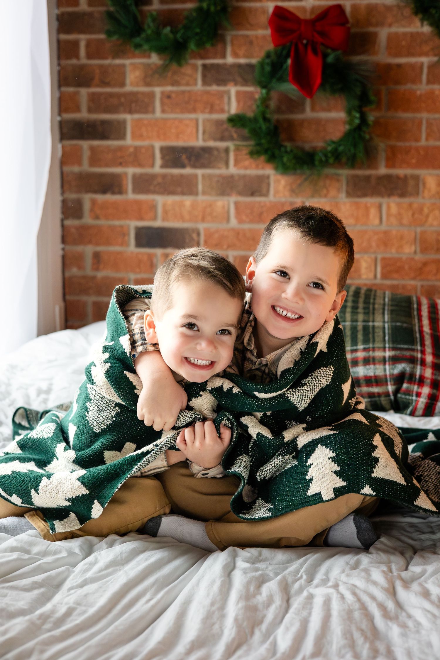 Siblings sharing joyful moments under a Christmas-themed blanket against brick wall.