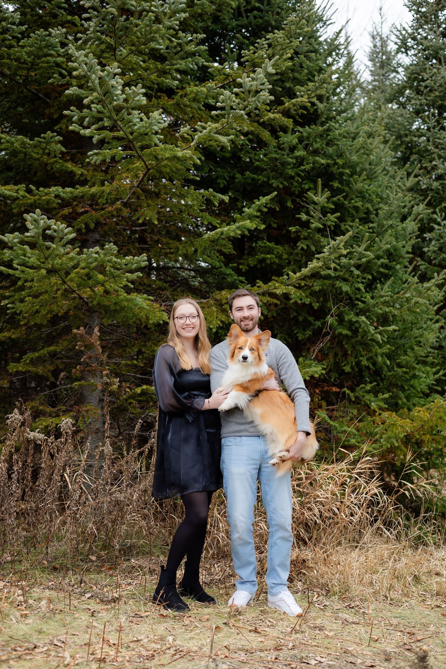 A couple poses with their corgi pet in a natural outdoor setting with pine trees.