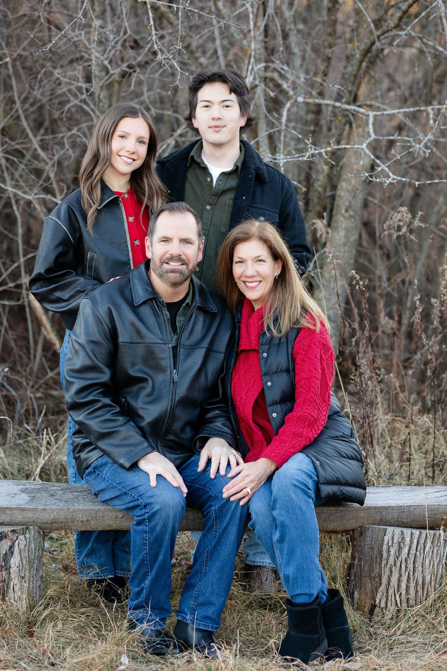 Casual outdoor family portrait on rustic wooden bench surrounded by bare winter trees.
