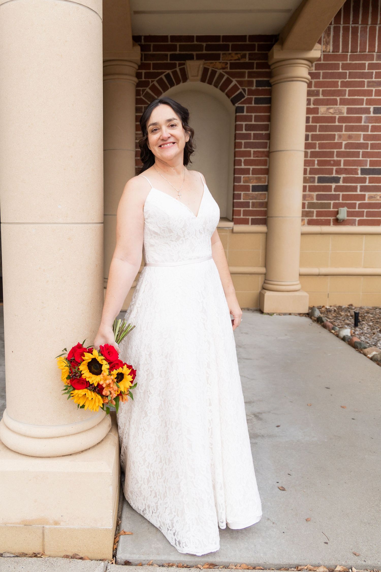A white lace wedding dress is showcased in front of tan columns and brick architecture.