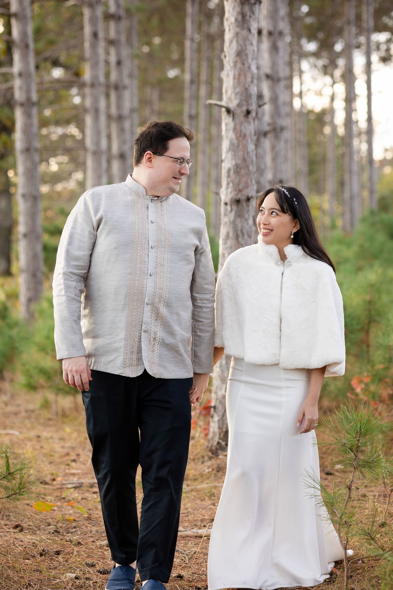 A couple in white and black attire share romantic moments while walking through a sunlit pine forest.