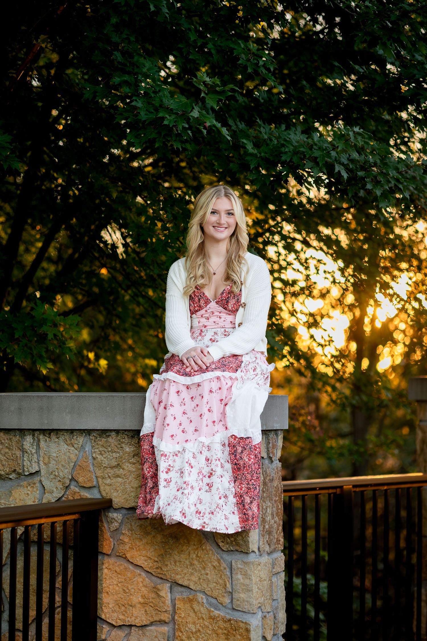 Woman in red and white dress poses by stone wall railing at sunset.