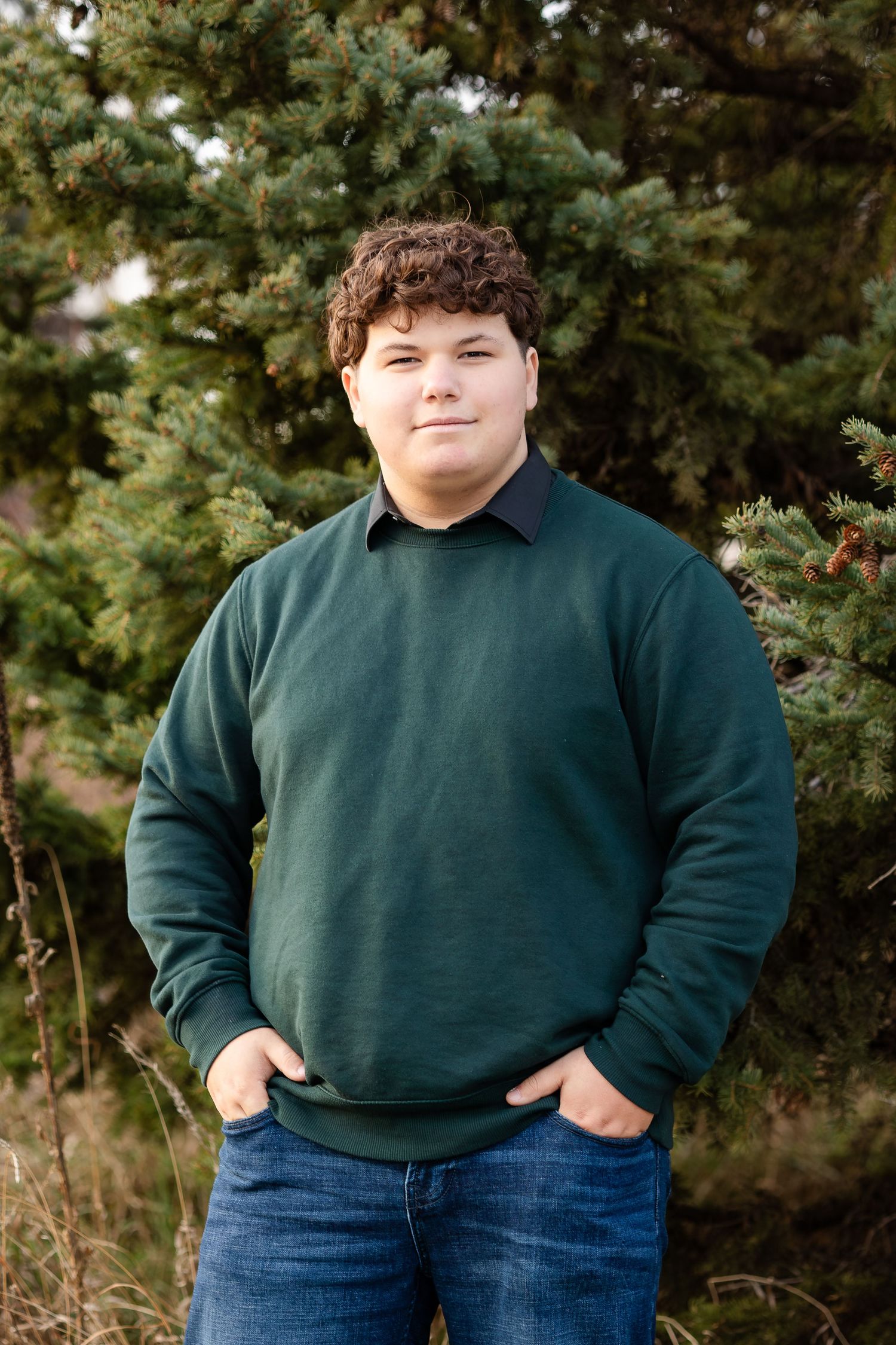 Person in green sweater and jeans stands with hands in pockets against evergreen trees.