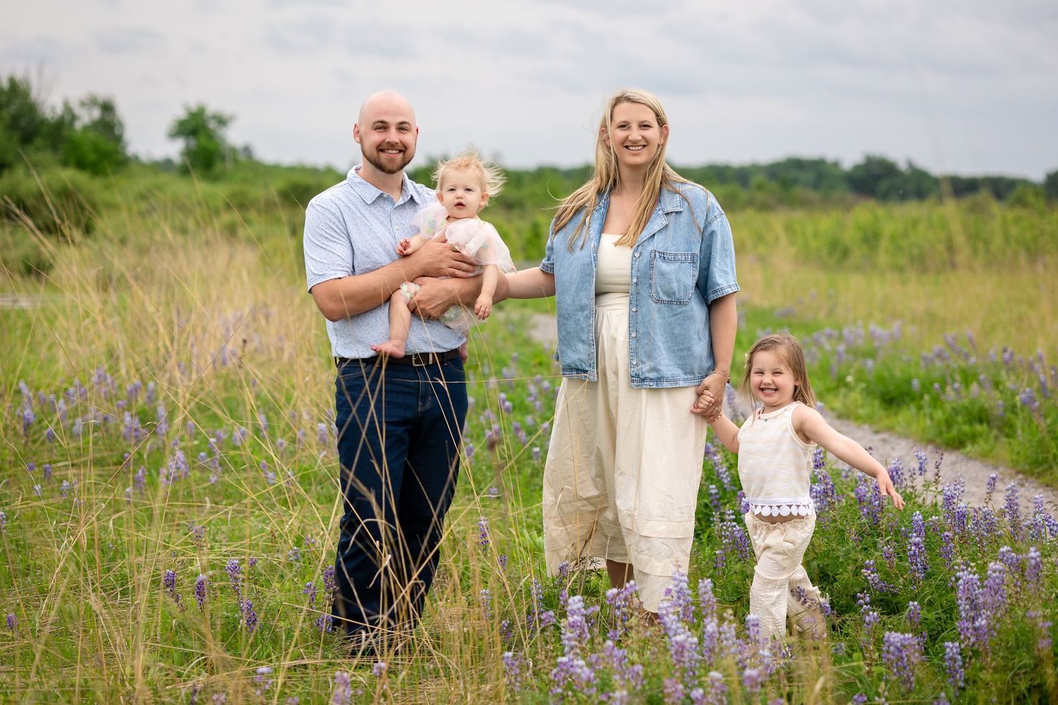 Family of four posing in a wildflower field, parents holding young children, smiling outdoors on a cloudy day.