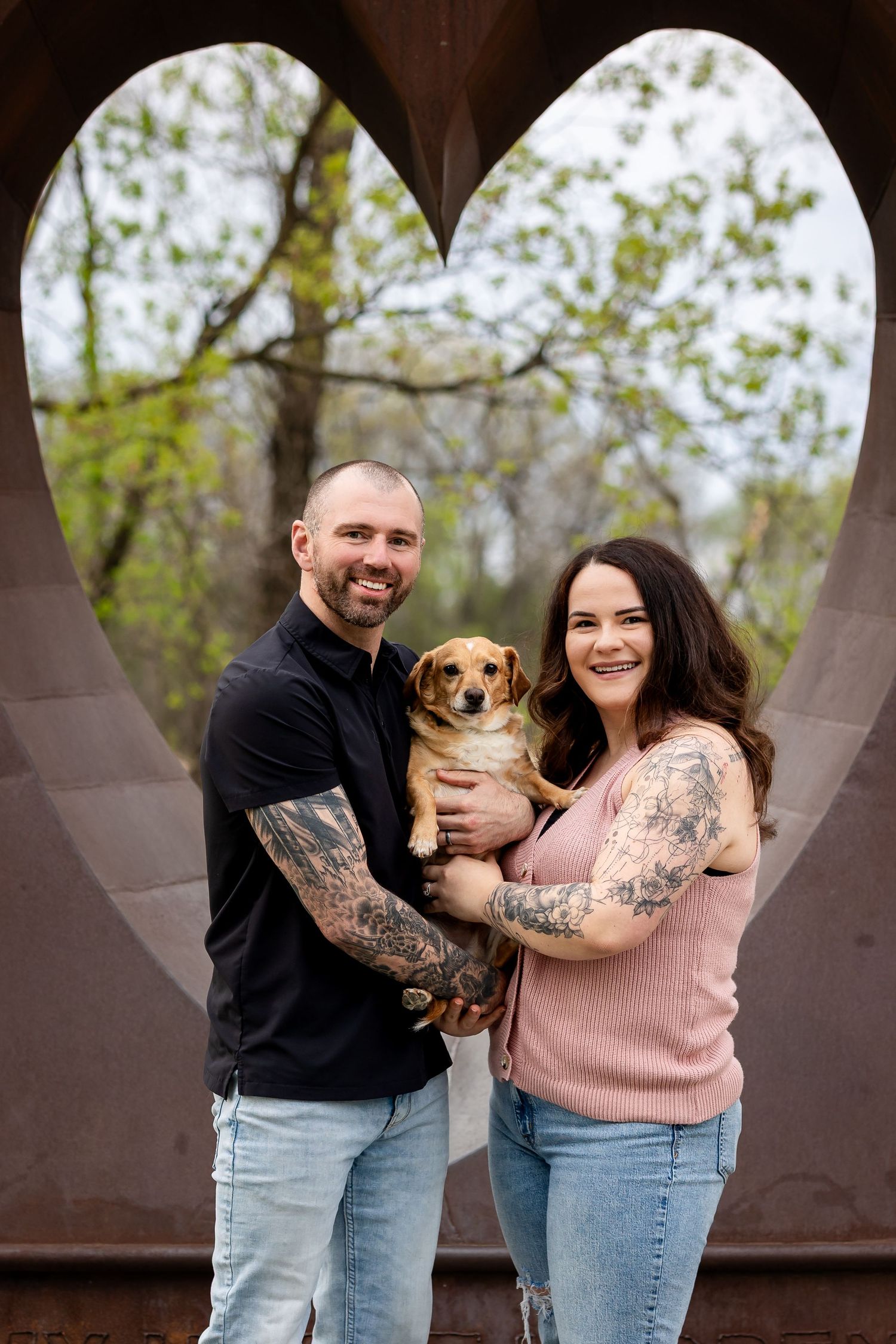 Smiling tattooed couple holding a small dog framed by a heart-shaped sculpture outdoors.